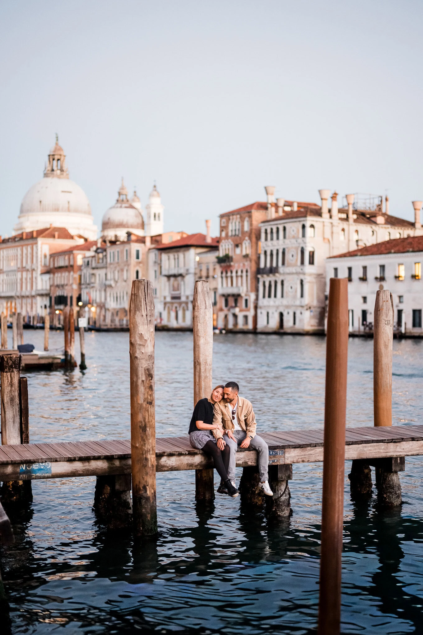 A couple sharing an intimate moment on a wooden dock in Venice at sunset, with historic buildings an.