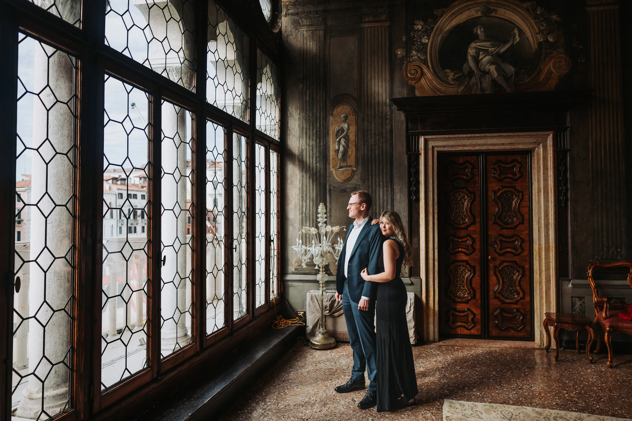 A man and woman stand together inside an ornate, historic building, gazing out through large, patter.