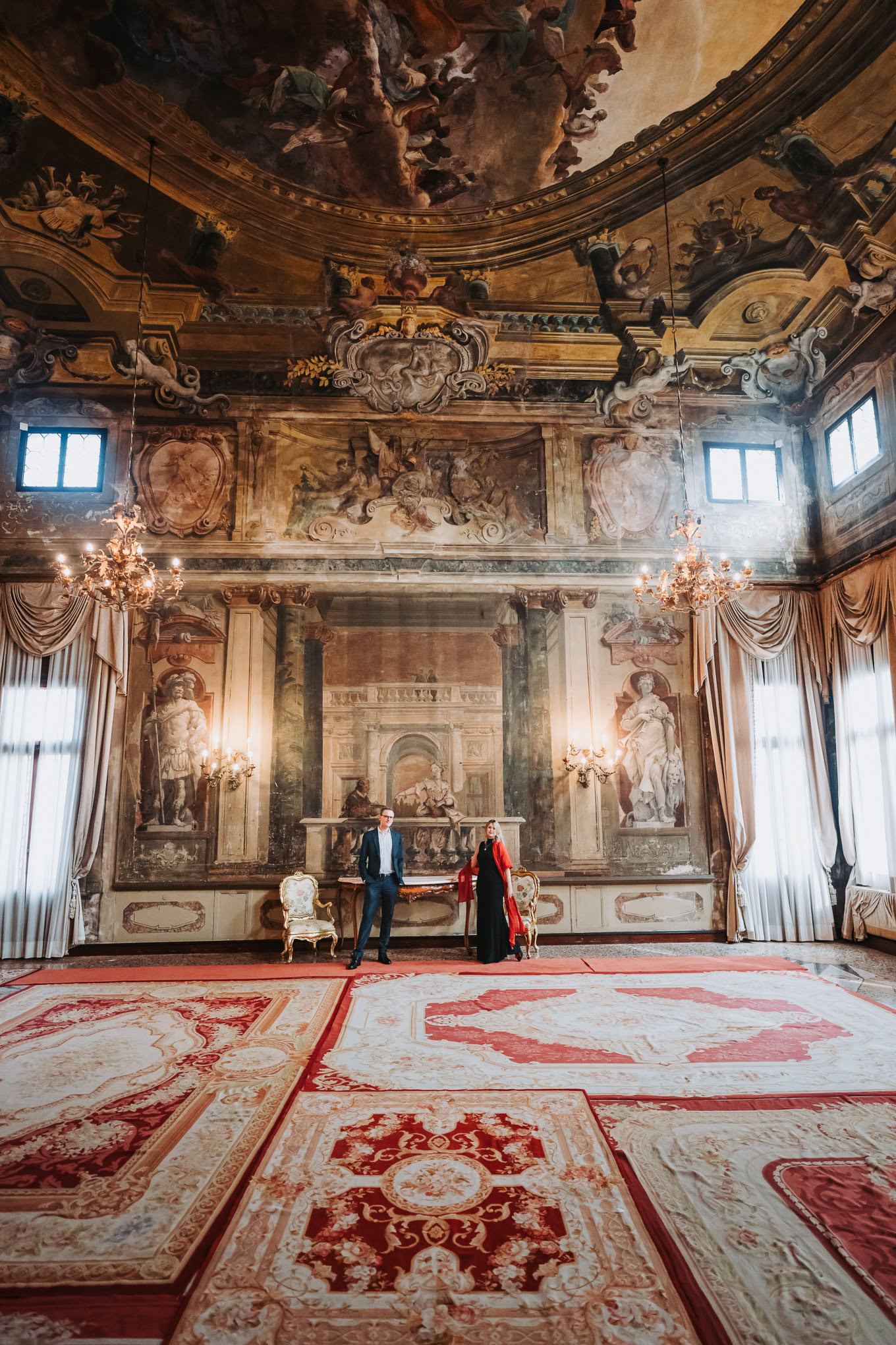 A woman and a man stand in an opulent, baroque-style room with elaborate frescoes, gilded details, a.