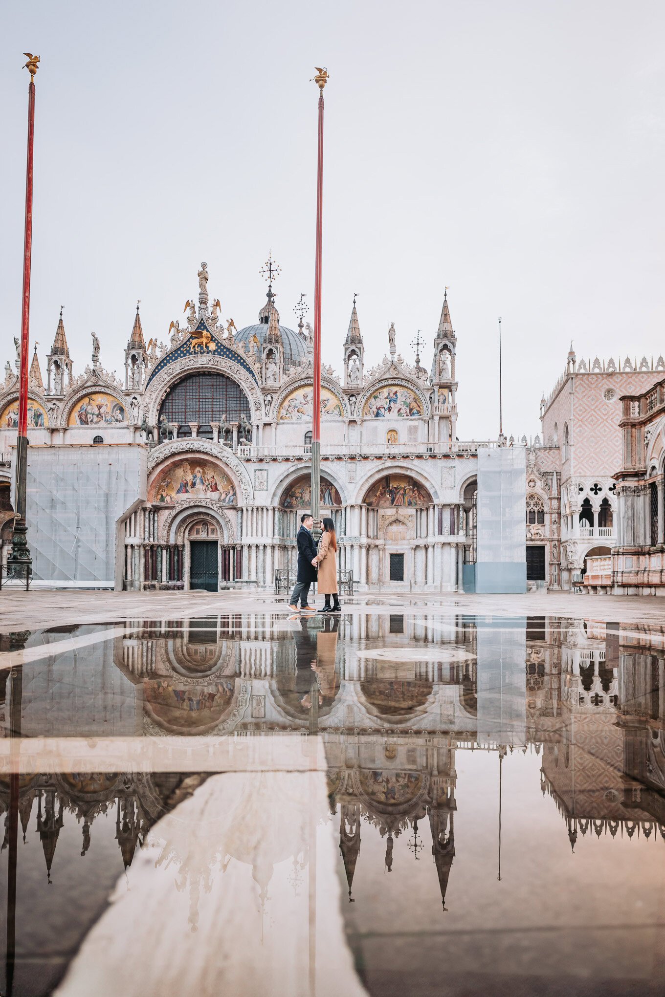 A couple stands close together in front of a grand, ornate cathedral on a rainy day, their reflectio.