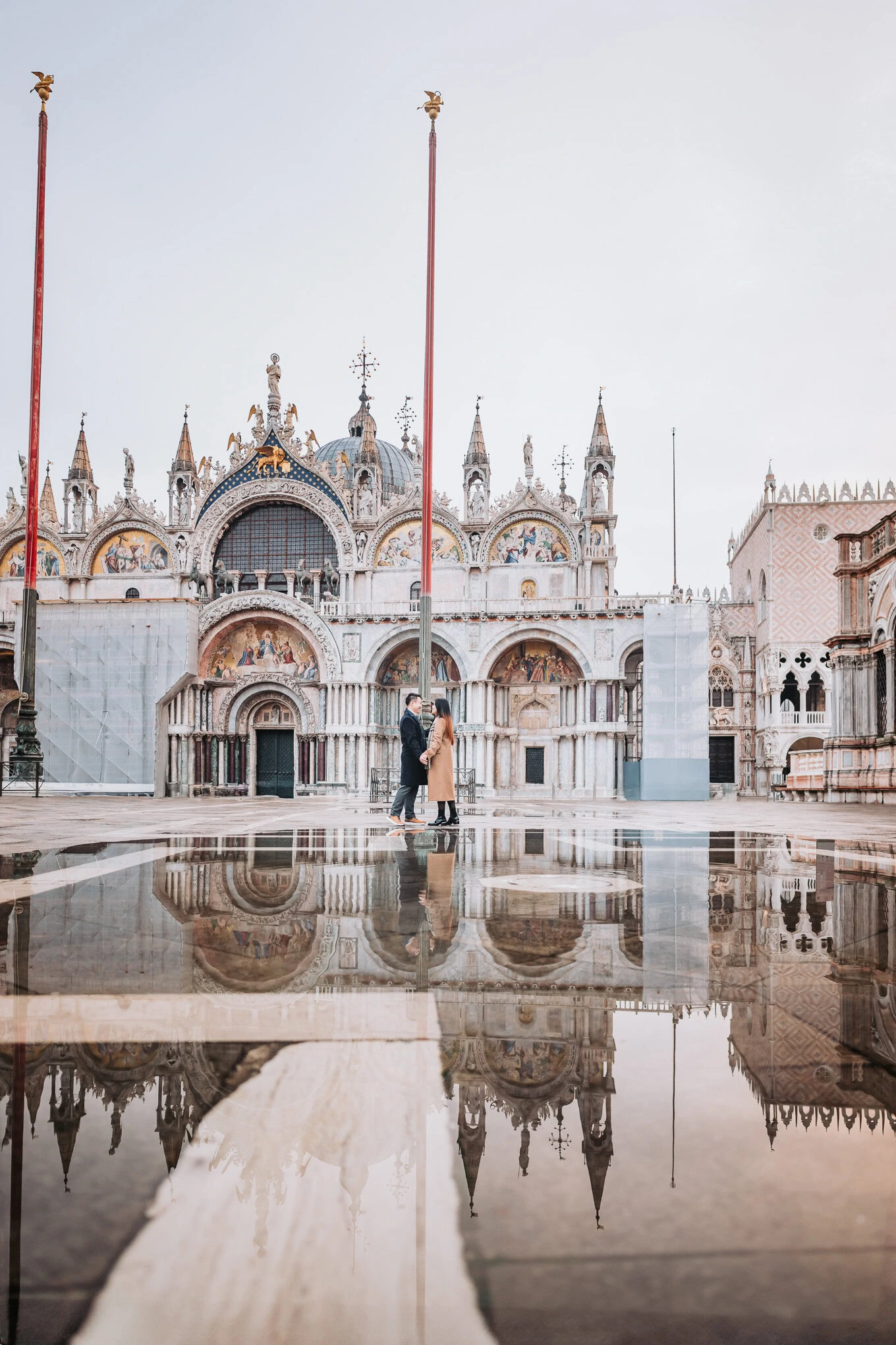 A couple stands close together in front of a grand, ornate cathedral on a rainy day, their reflectio.