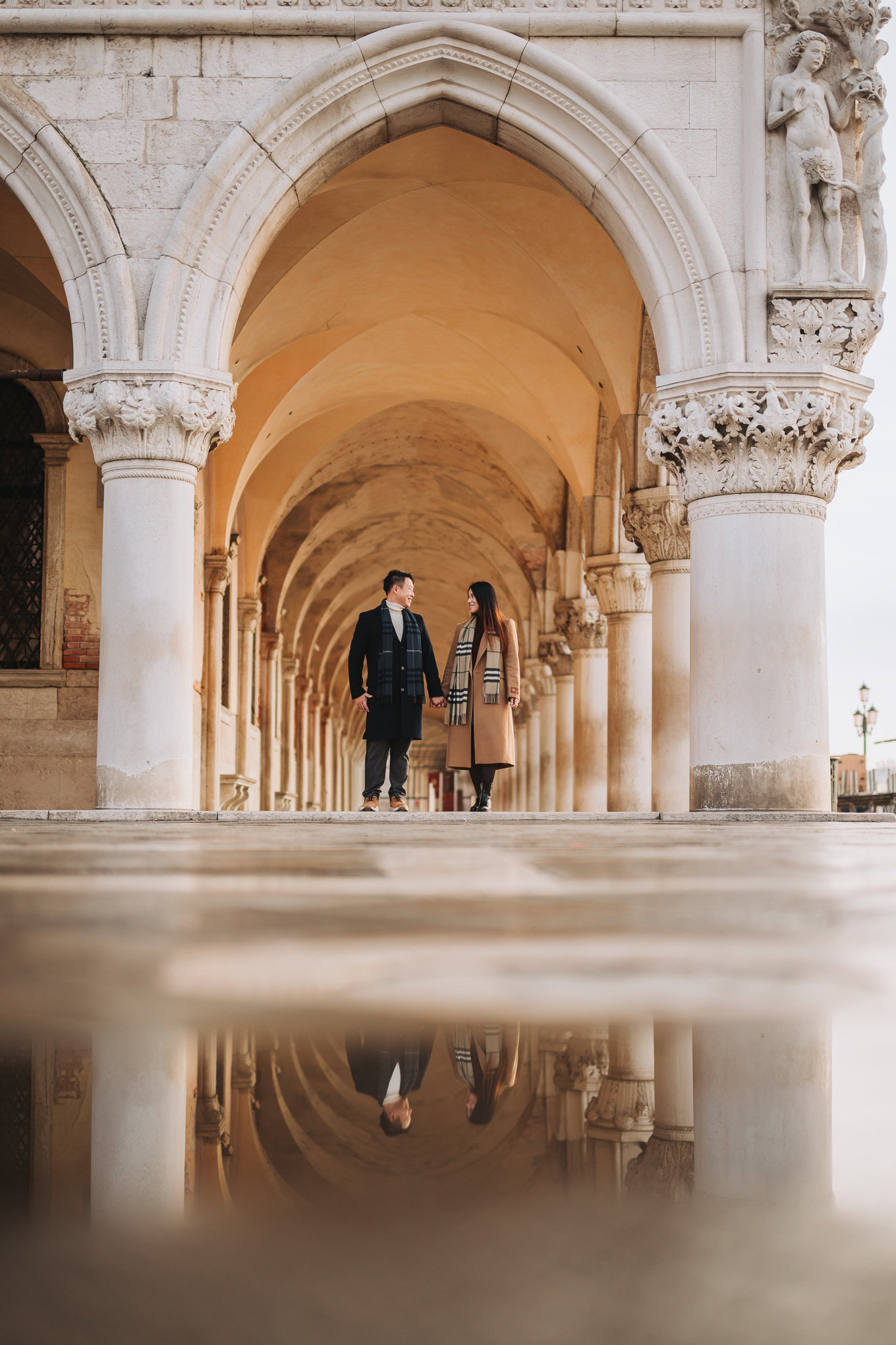 Two people walk beneath historic stone arches at dusk, illuminated by soft warm light, creating a pe.