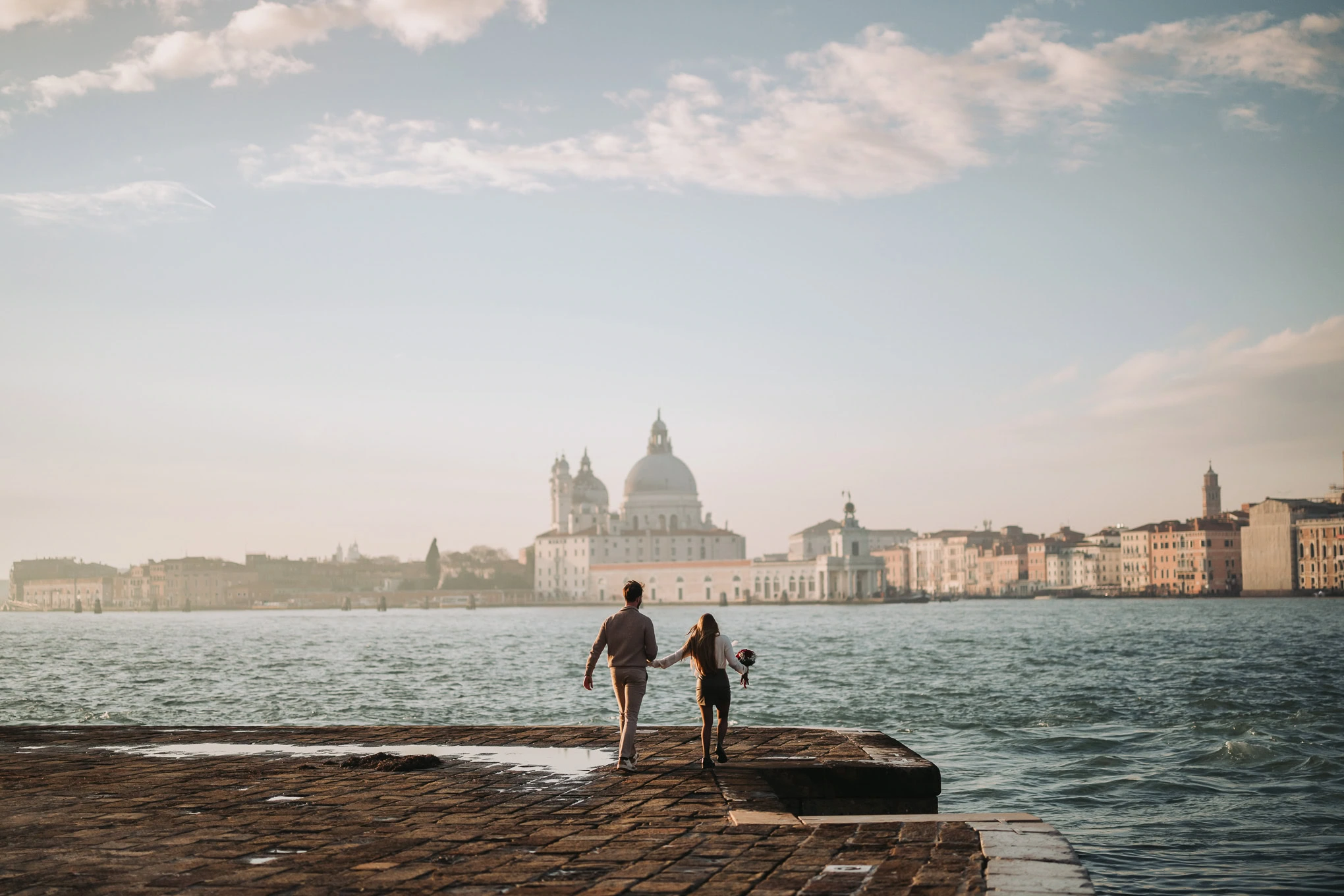 A couple walking hand in hand along a waterfront promenade at sunset, with a historic cityscape and.