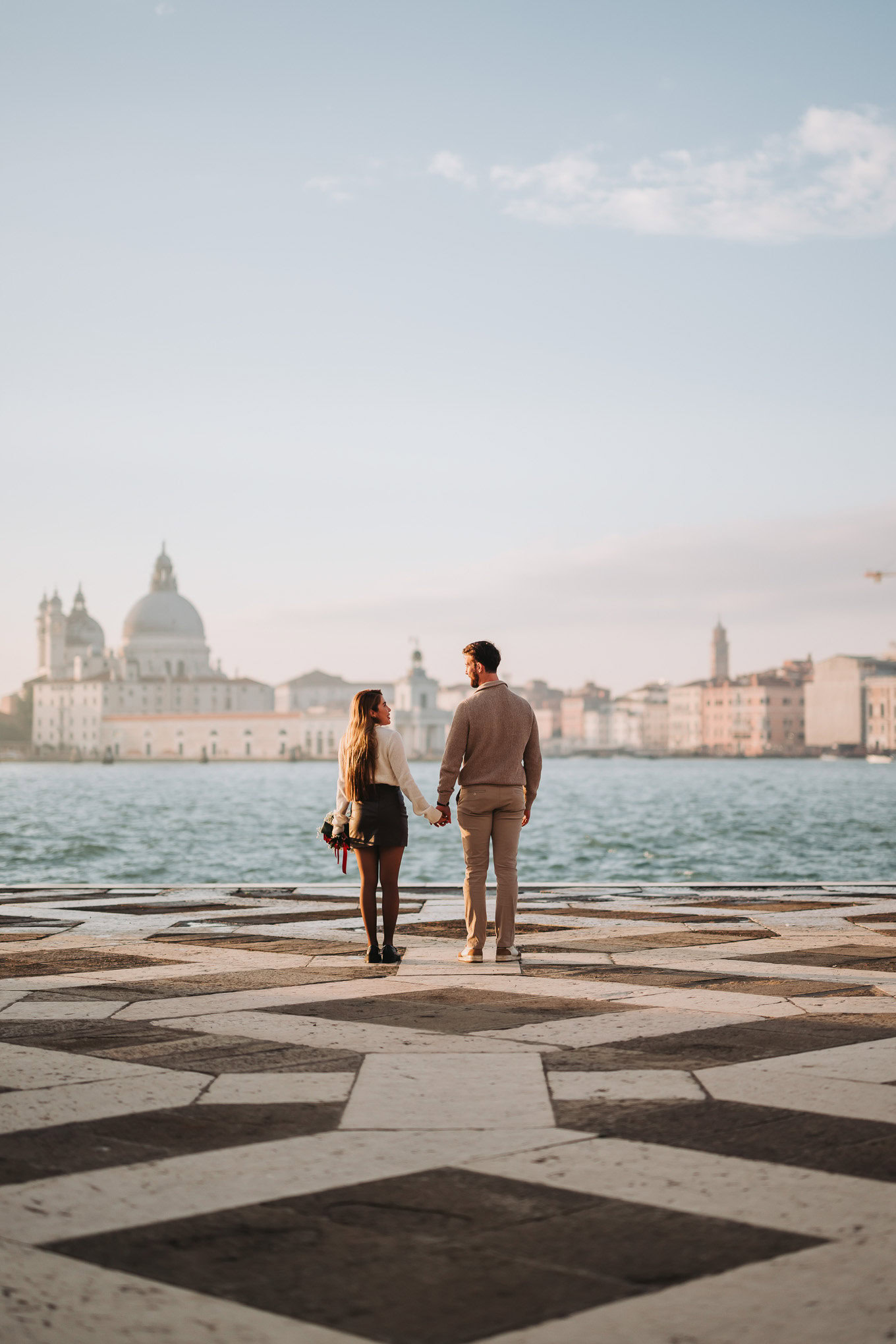 A couple holding hands and gazing across the water at sunset, with a historic cityscape in the backg.