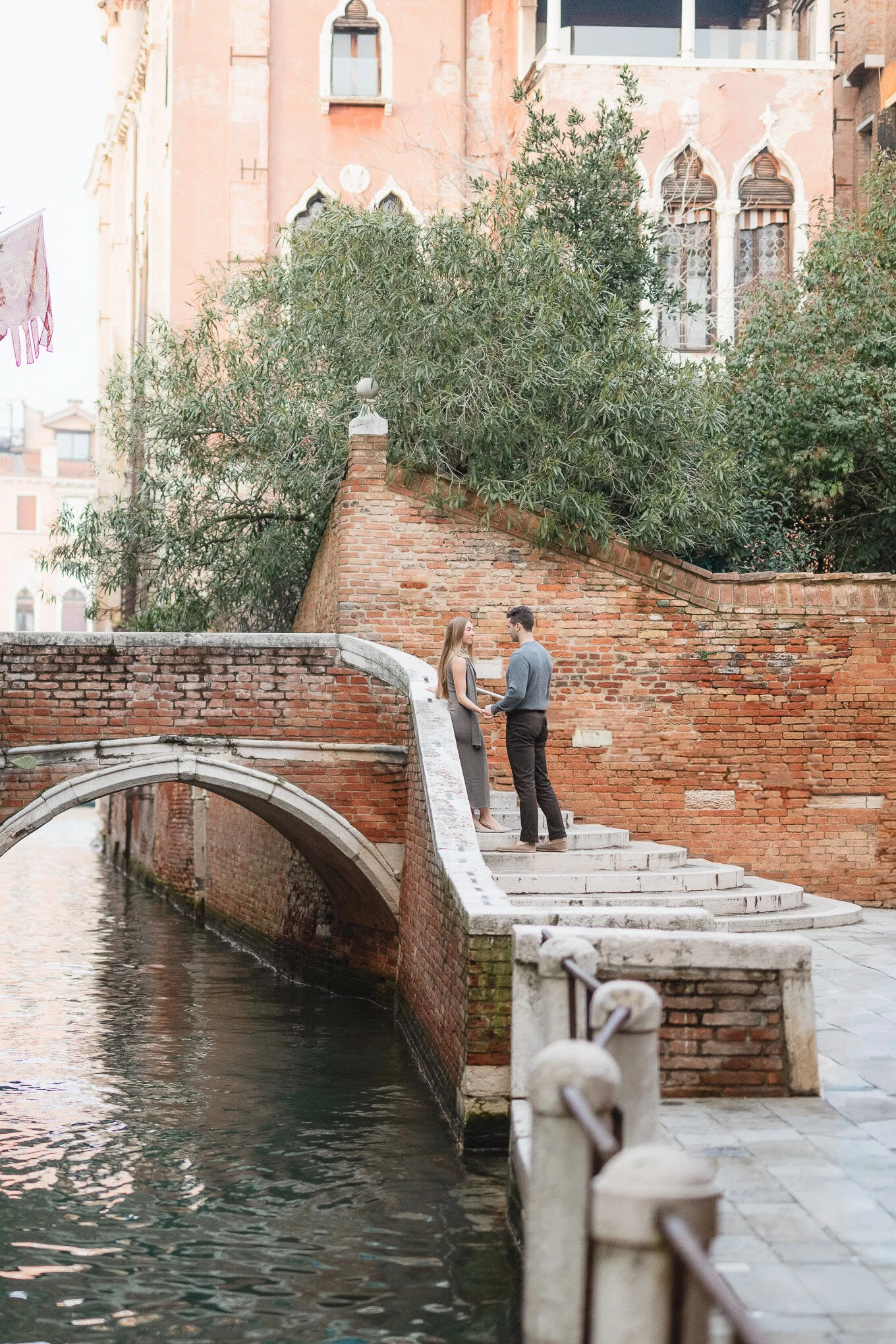 A couple stands on a small bridge over a canal in Venice, surrounded by lush greenery and historic a.