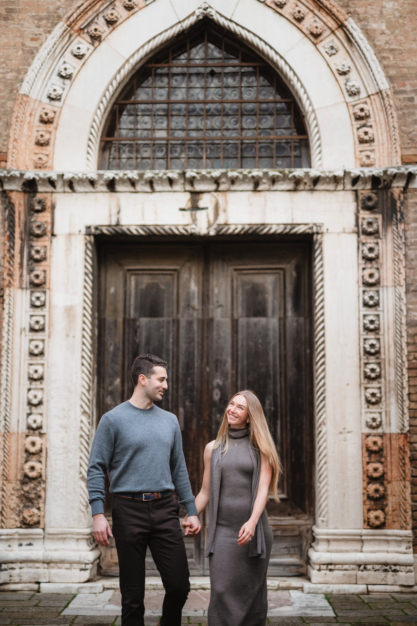 A couple holding hands and smiling at each other in front of an ornate, historic church entrance bat.
