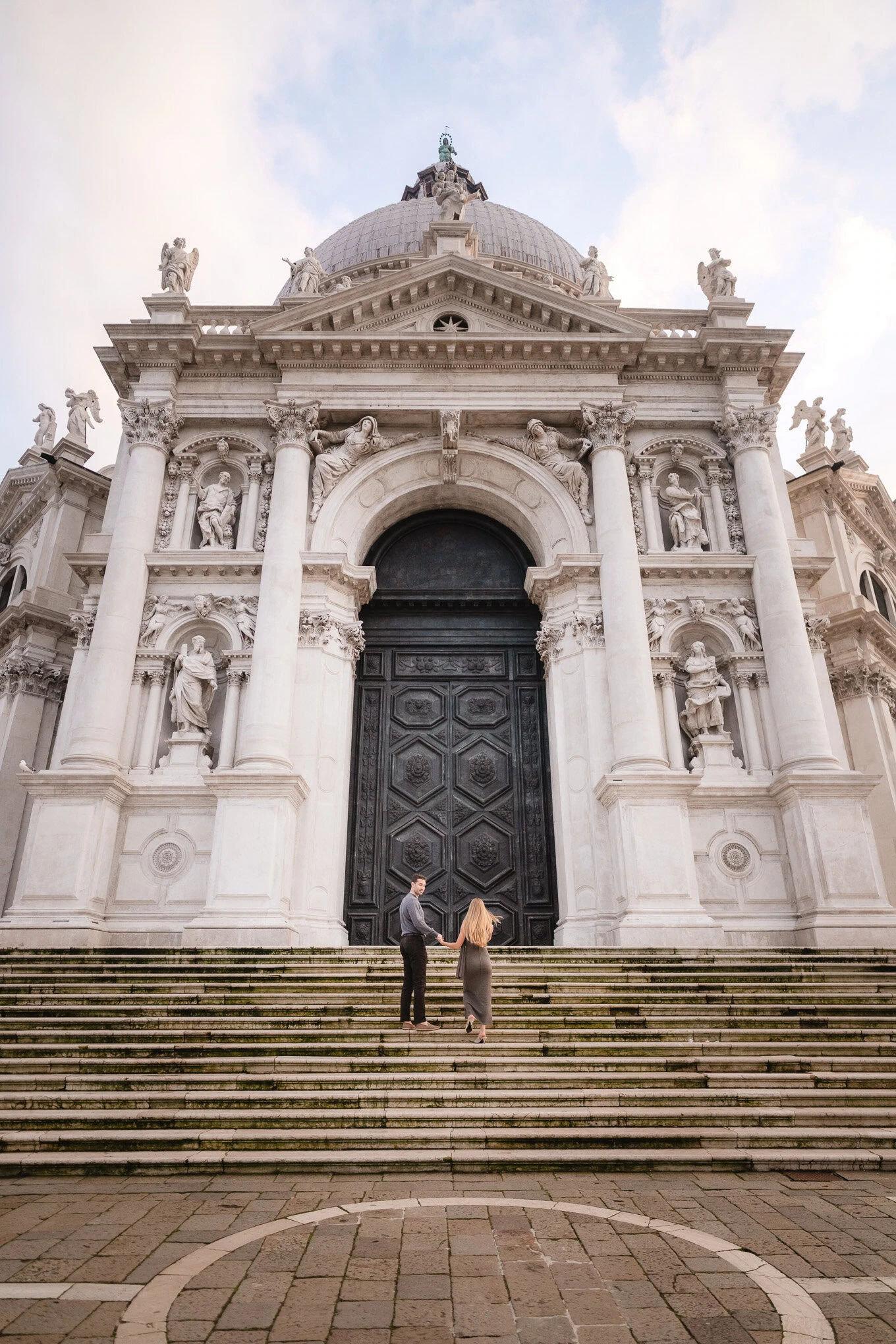 A couple holding hands and gazing at a grand historic cathedral under a softly lit sky.