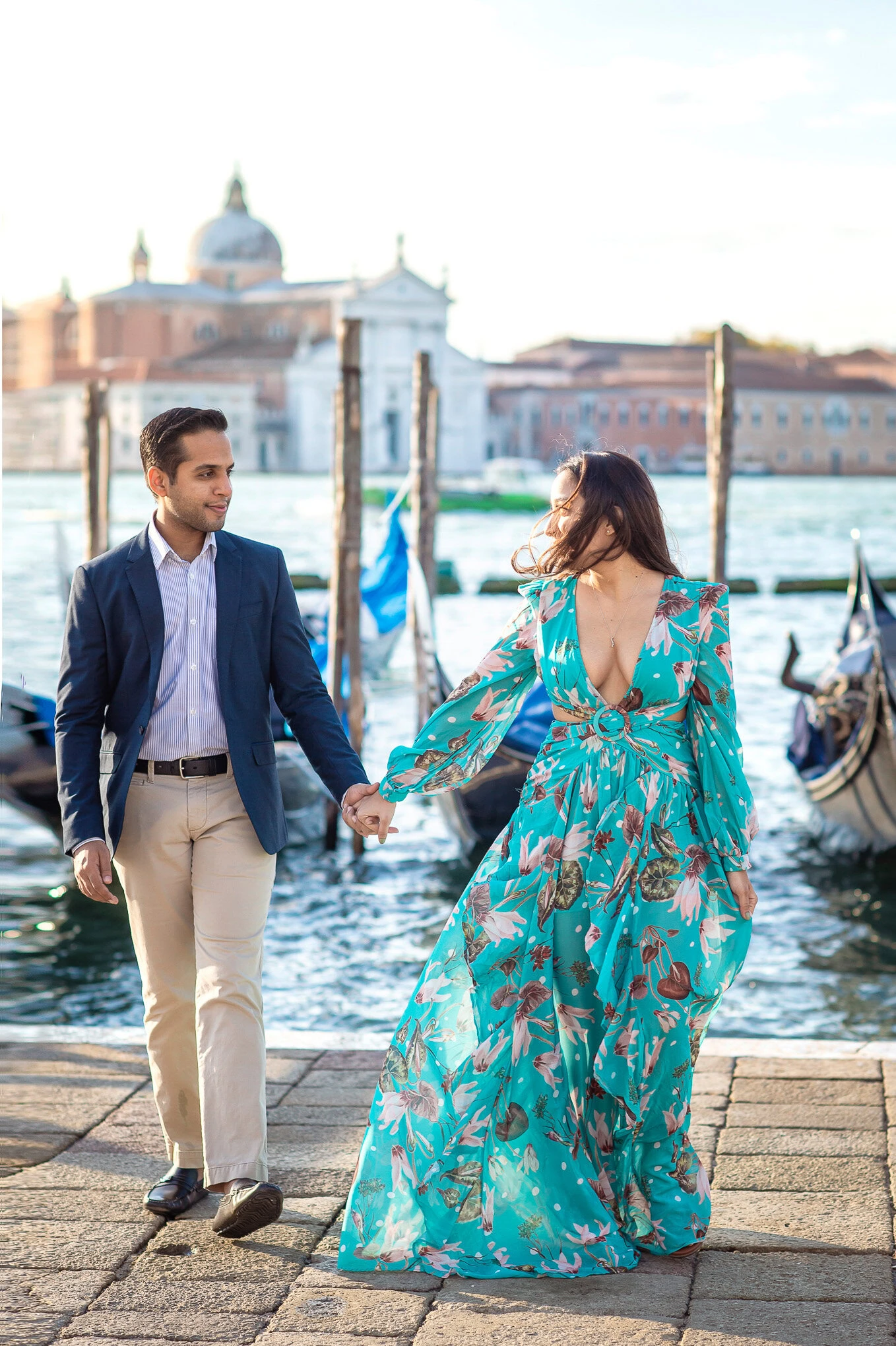 A woman in a flowing turquoise dress and a man in a navy blazer walk hand in hand along a Venetian c.