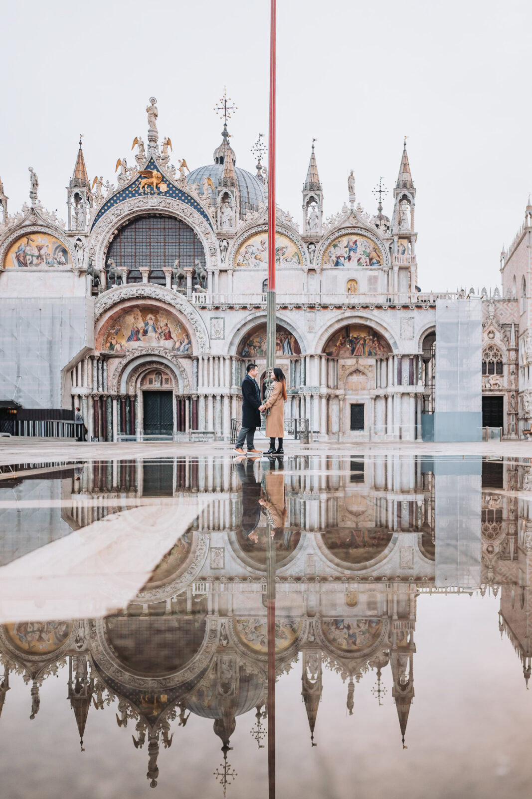 Flooded Piazza San Marco at sunrise during acqua alta, with two people sharing a moment amidst histo.