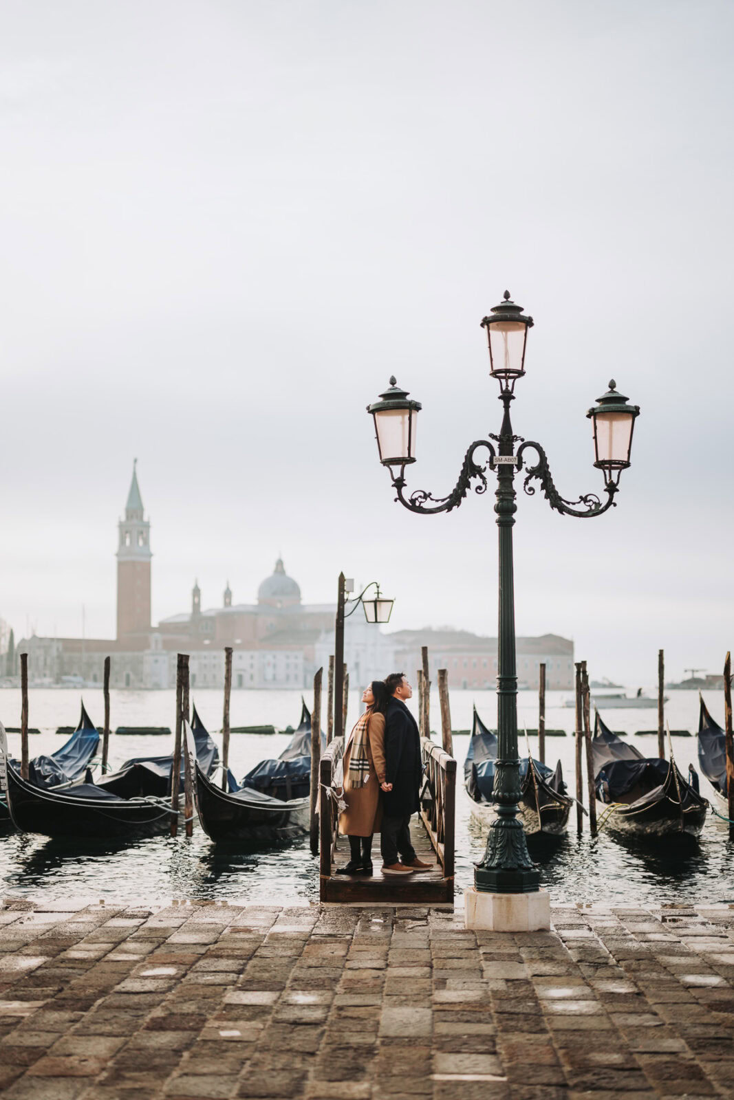 Venice gondola dock during acqua alta at sunrise with couple and historic cityscape in background.