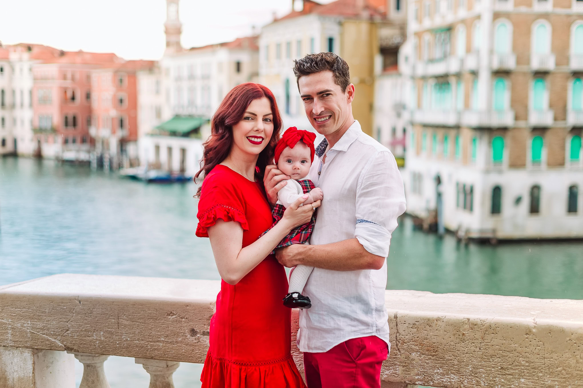 A woman, man, and baby enjoy a sunny day on a Venice canal bridge, surrounded by colorful historic buildings and gentle.