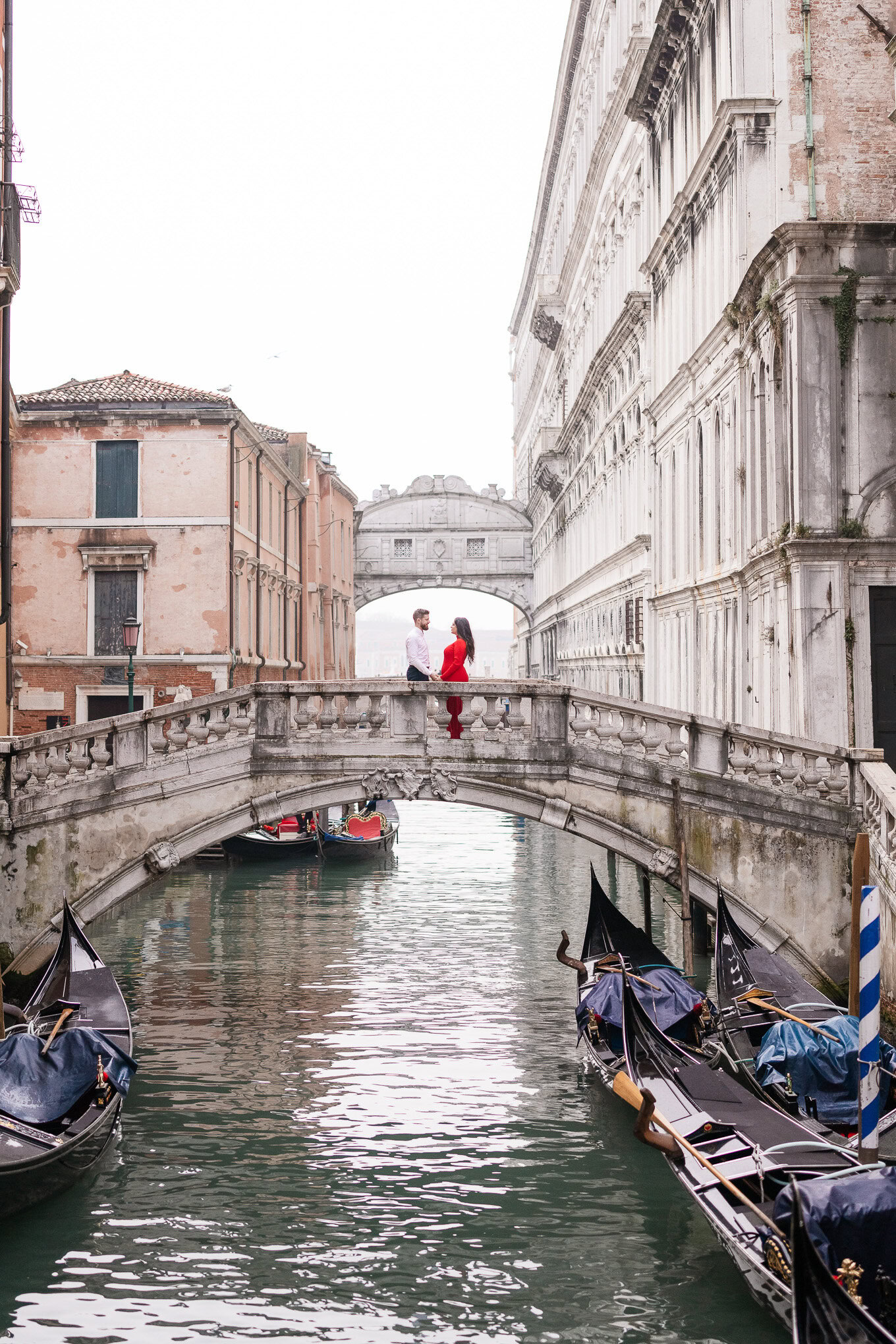 A family walks hand-in-hand across a small stone bridge over a Venetian canal, surrounded by historic buildings and gond.