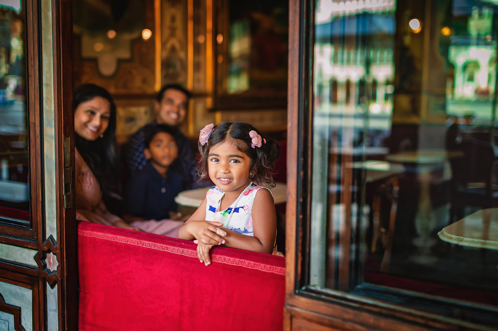 A girl with pigtails sitting at a Venetian café table, smiling warmly in a lively, atmospheric setting.