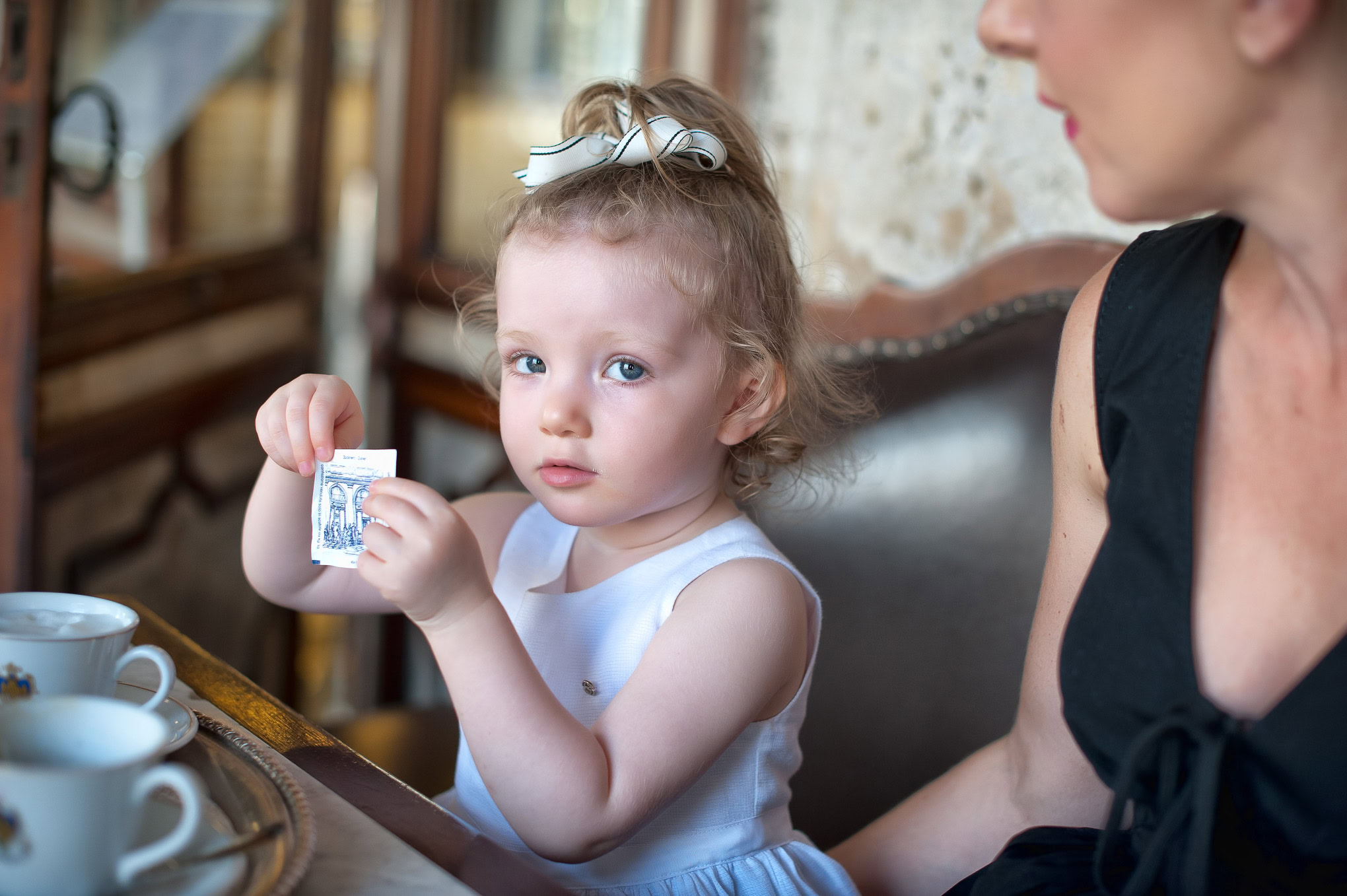 A young girl with curly hair and a white bow, sitting at a table with her mother, holding a small card in a cozy Venetia.