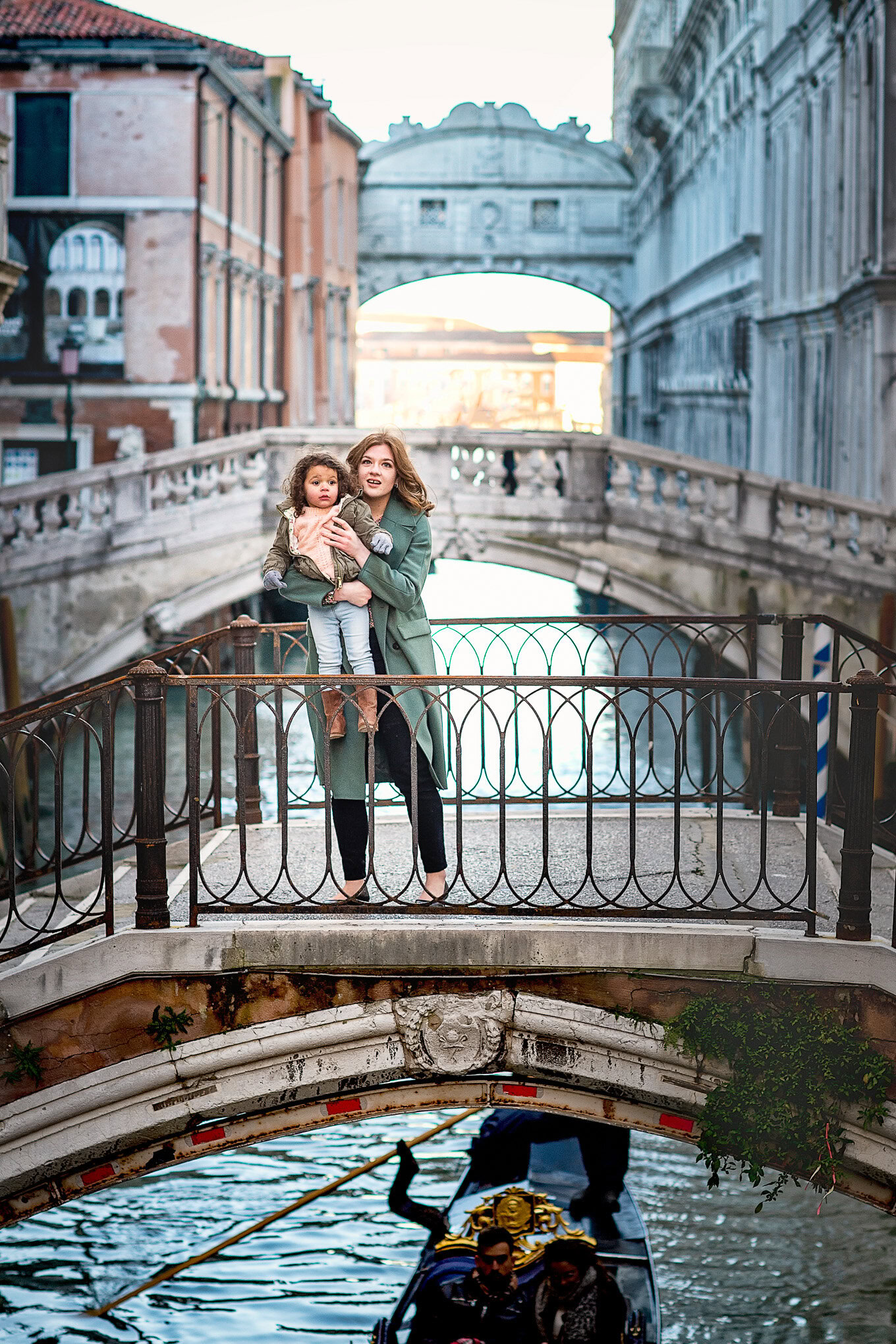 Two young girls sharing a moment on a small bridge over a Venetian canal, surrounded by historic buildings and soft even.