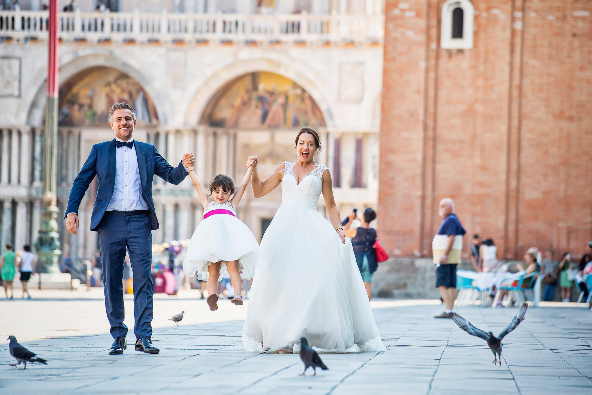 A woman and man in formal attire hold hands with a young girl in a white dress, joyfully swinging her between them in a.