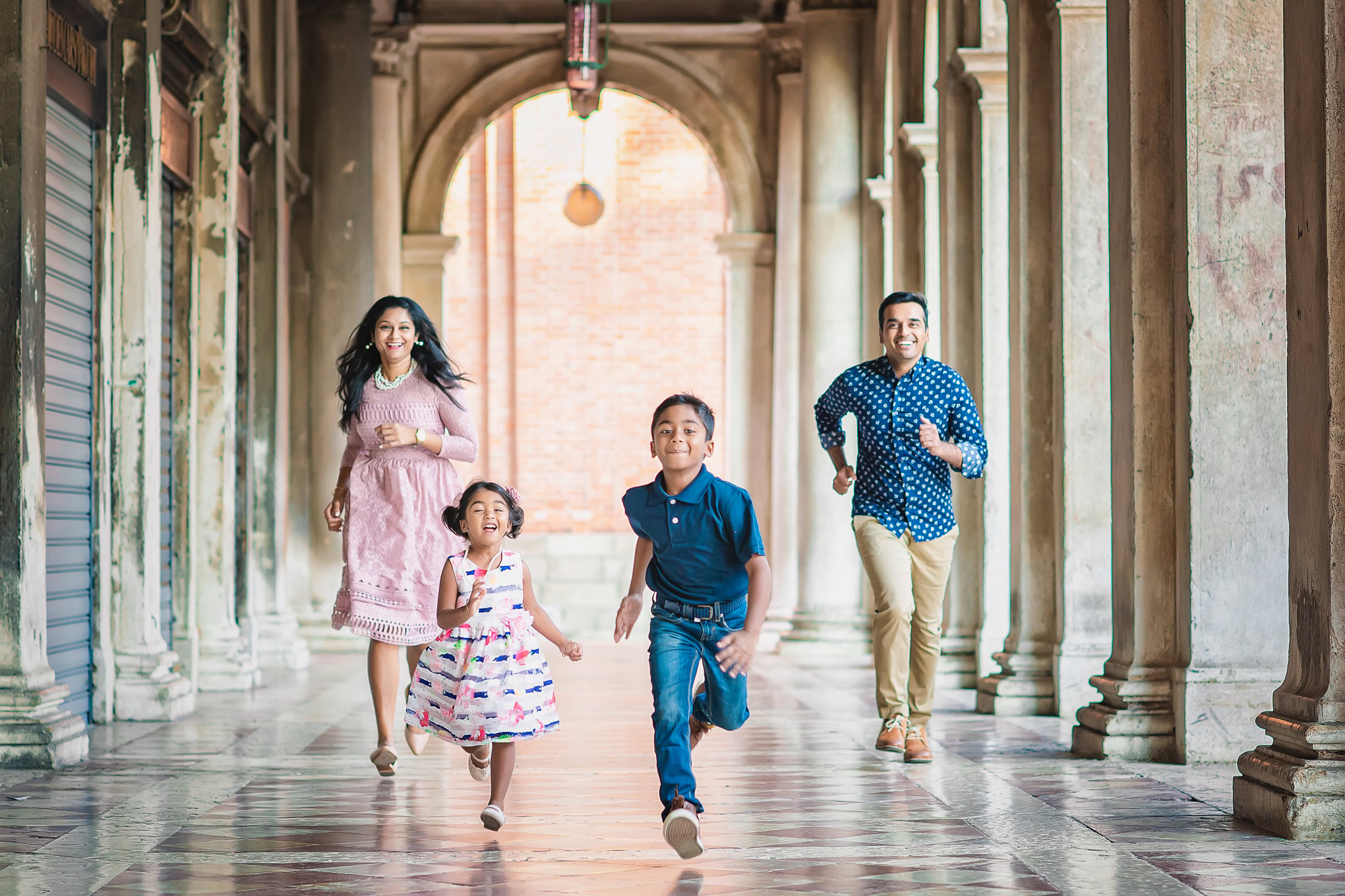 A family of four running and playing together through a historic Venetian arcade with warm natural light.