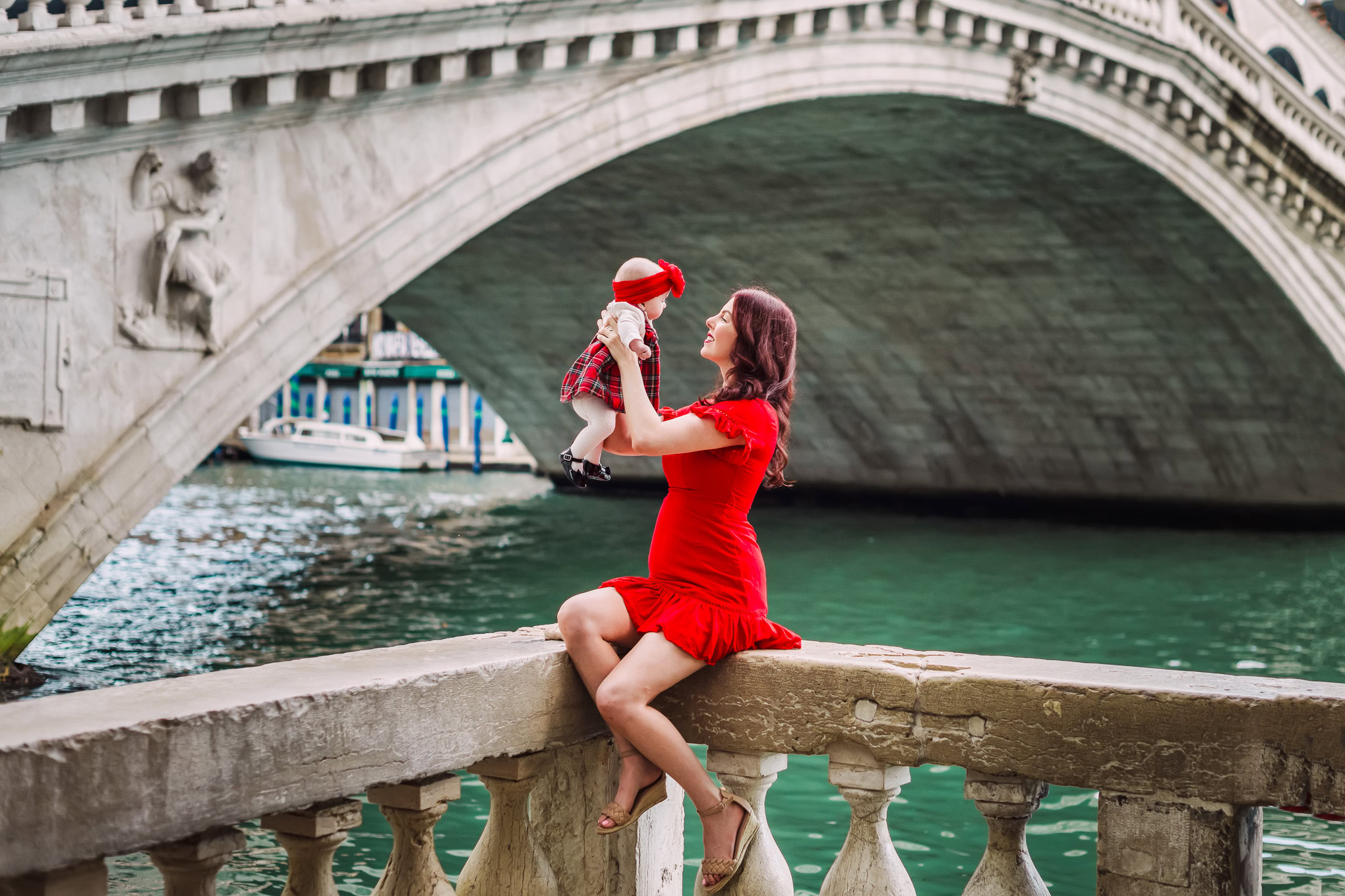 A woman in a red dress holding a child in a red hat by the Venetian canal under a historic bridge.