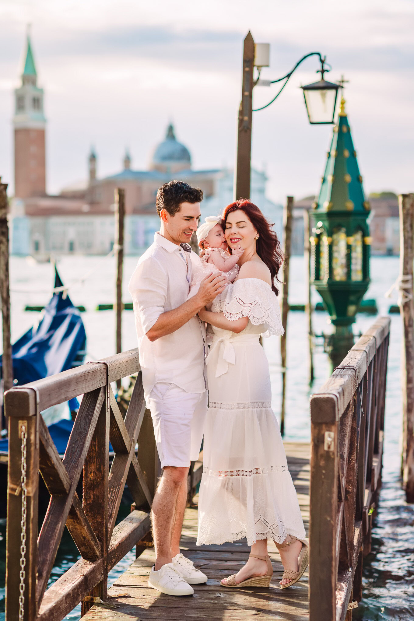 Family enjoying a tender moment on a Venice dock with historic buildings and water in the background, bathed in soft nat.