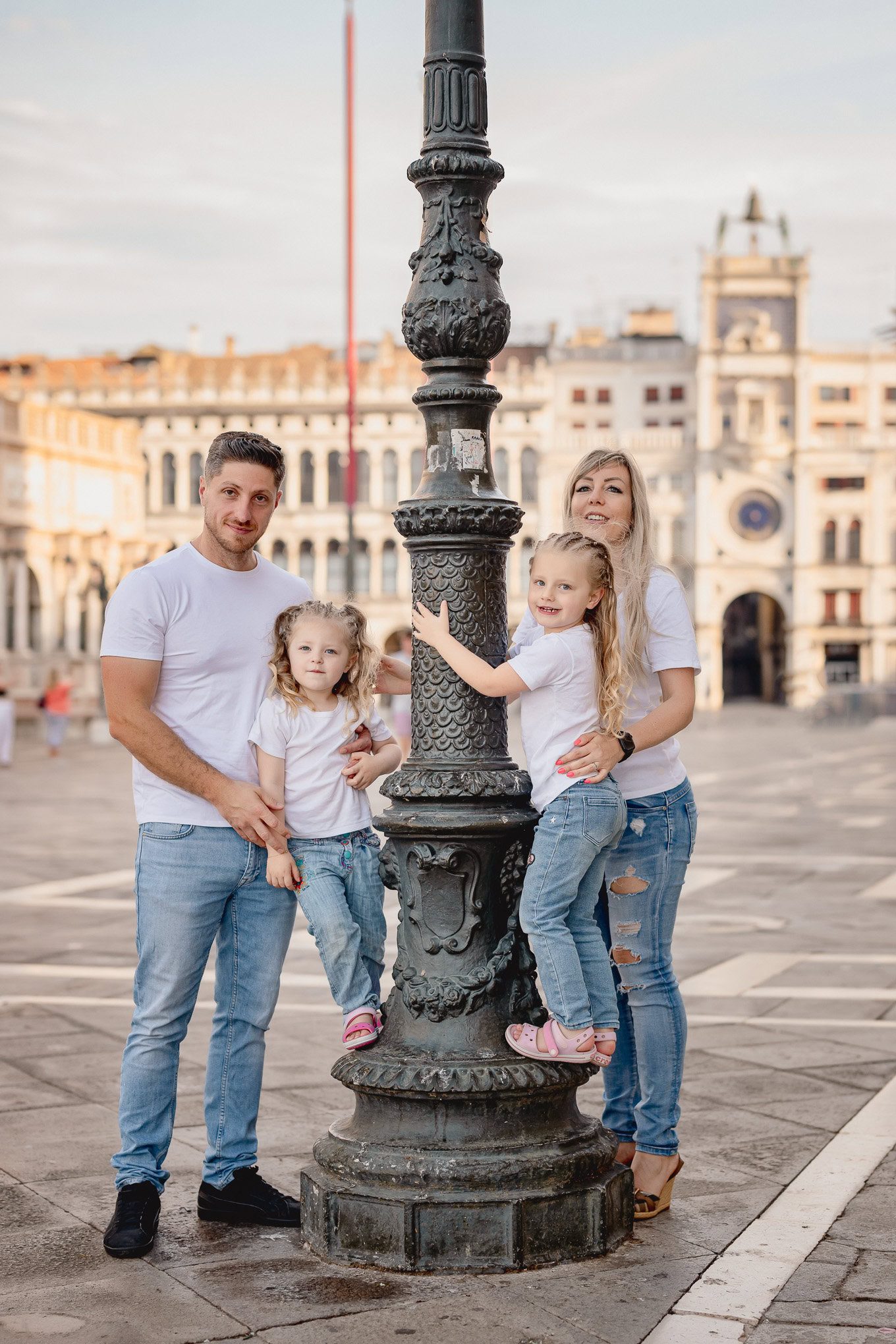 A family of four enjoying a playful moment around a historic lamp post in Venice's lively square during daylight.