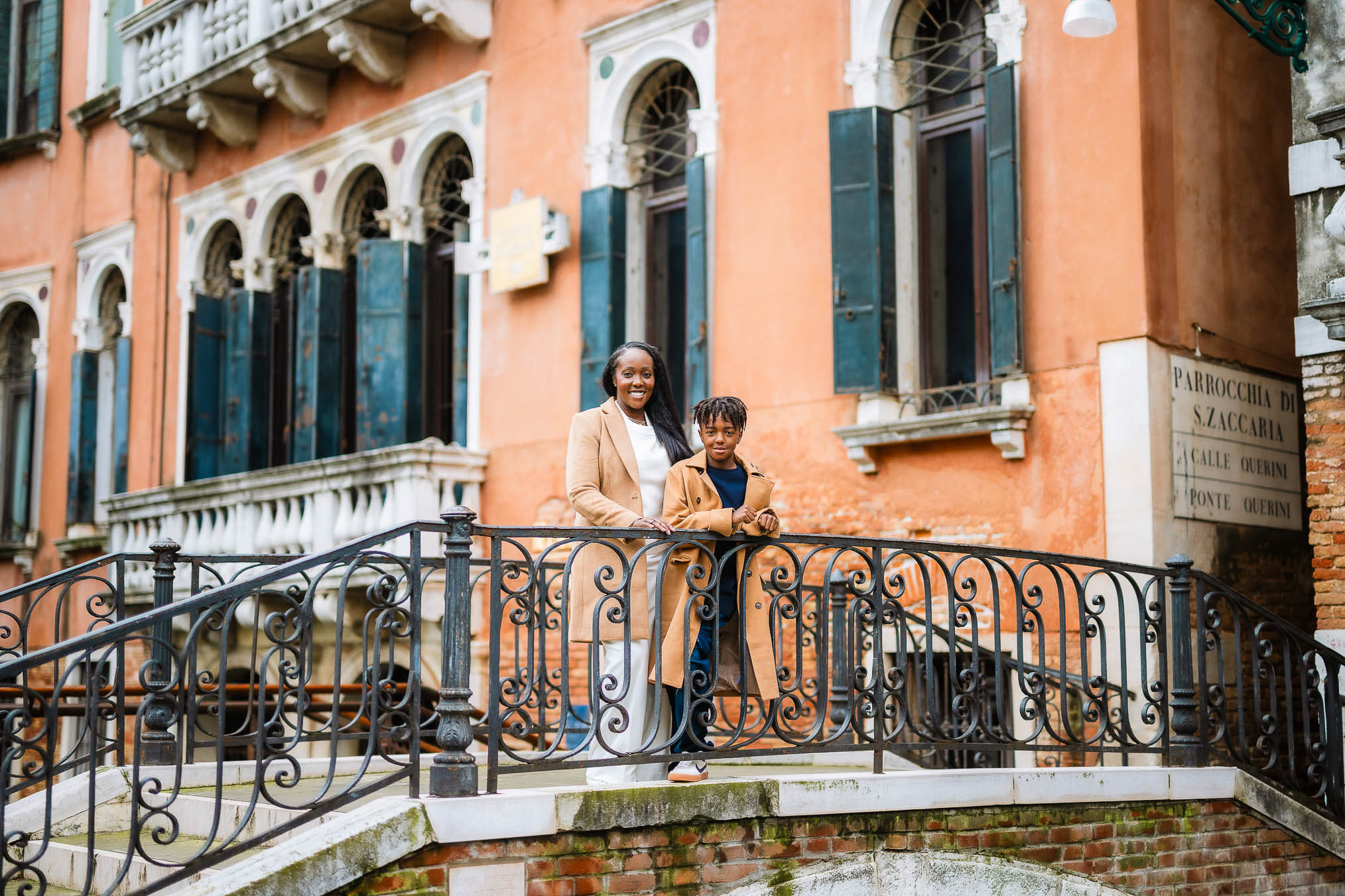 A woman and a boy stand on a decorative bridge in Venice, enjoying a sunny day with historic buildings in the background.