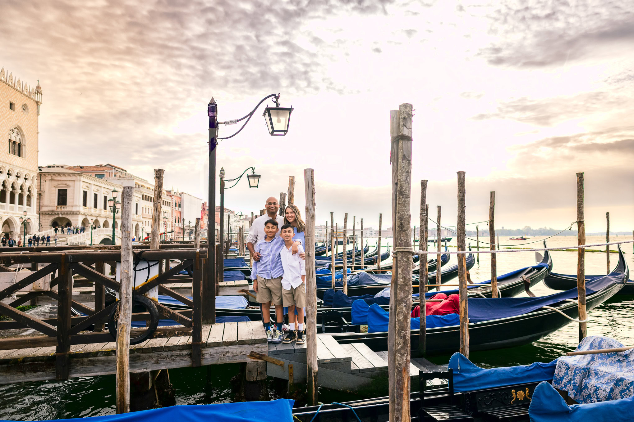 A family enjoying a walk along a Venetian dock with boats, under a cloudy sky, capturing a moment of togetherness and ex.