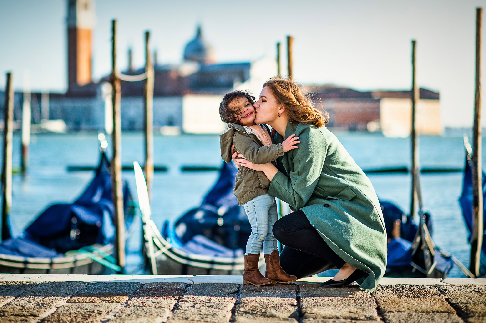 A woman and a young girl share a joyful kiss on a Venice canal promenade, with gondolas and historic buildings in the ba.
