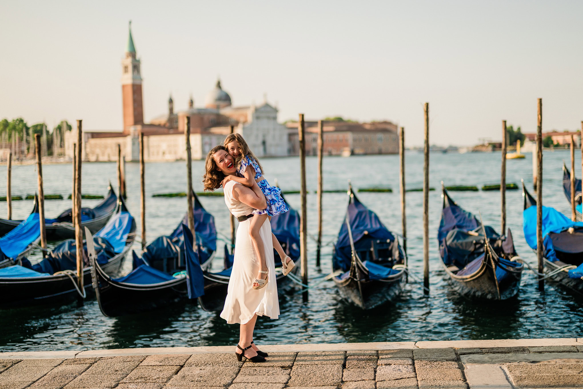 A woman holding a child by the Venetian canal with gondolas and historic buildings in the background.