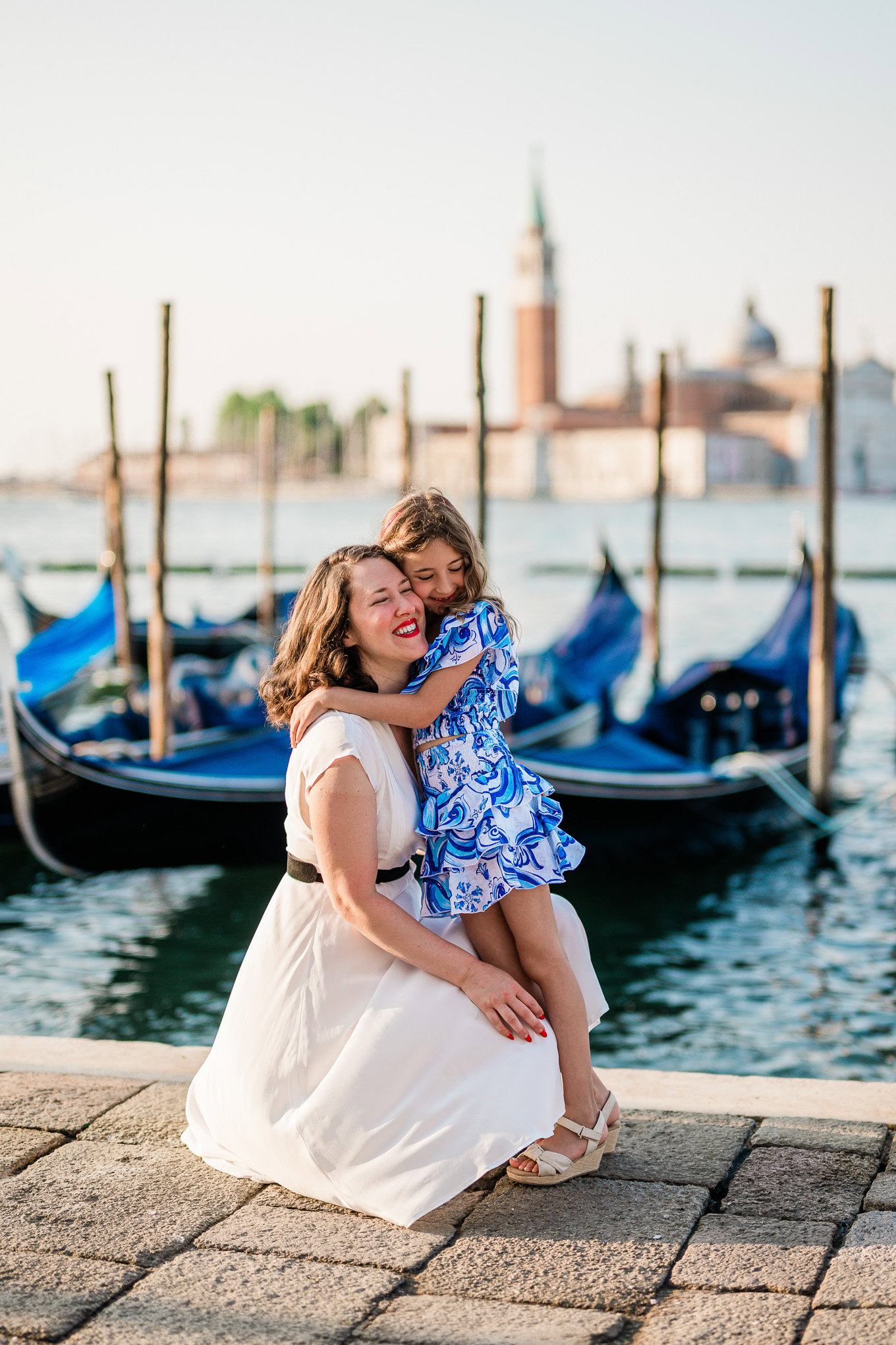 A woman and a young girl share a joyful moment by the Venice waterfront, with gondolas and historic buildings in the bac.