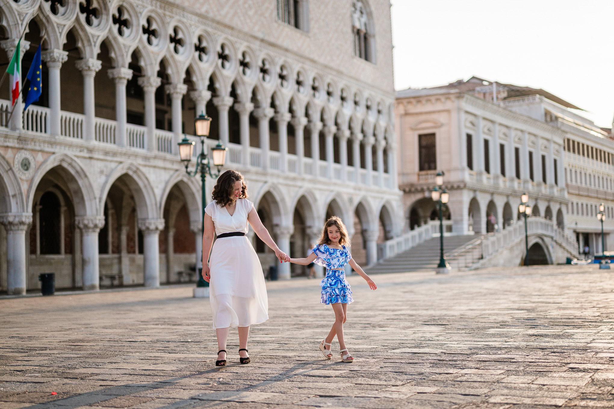 A mother and daughter holding hands and walking together through a historic Venetian square in natural daylight.