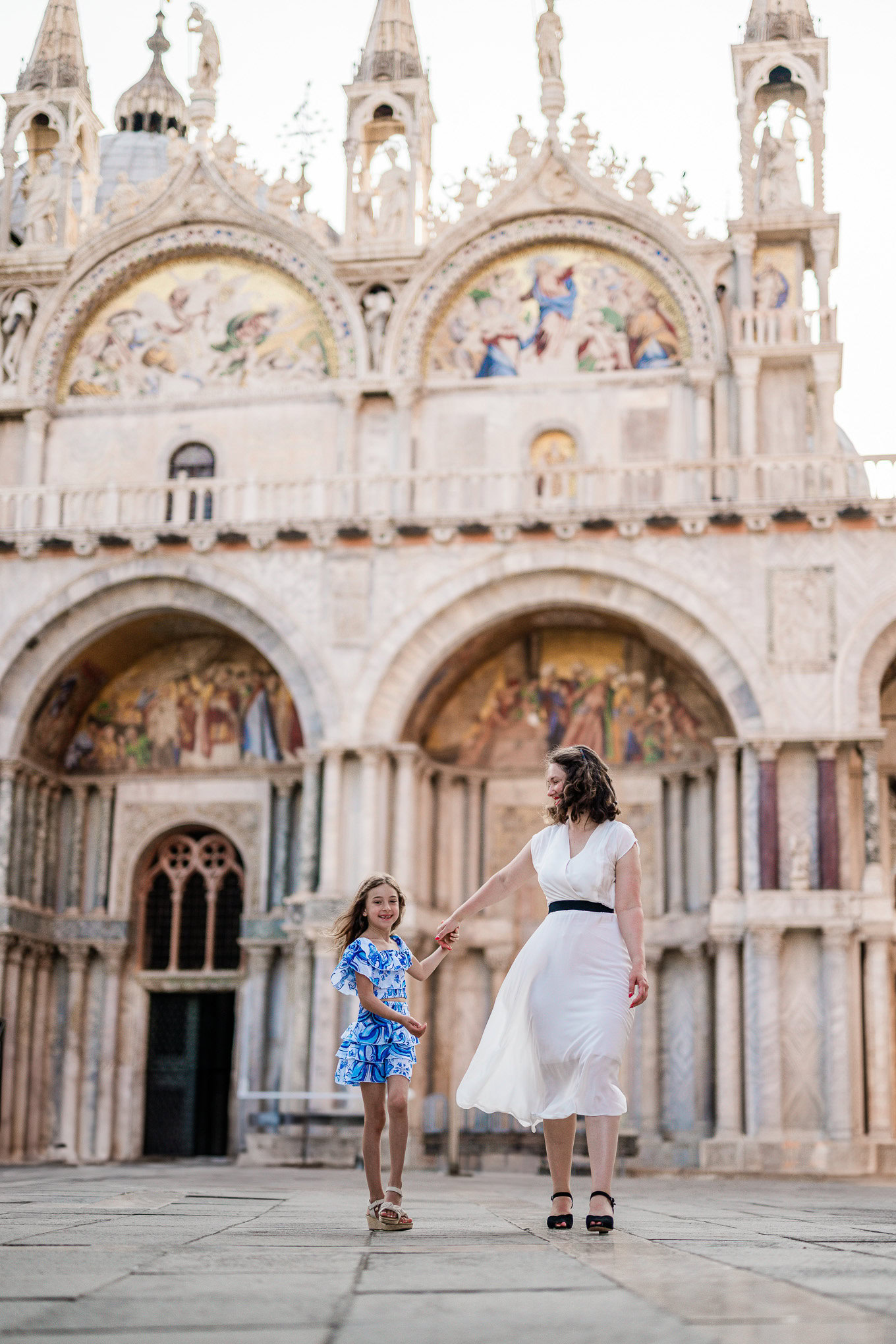 A woman and girl holding hands and walking together in front of a historic Venetian cathedral, bathed in soft natural li.