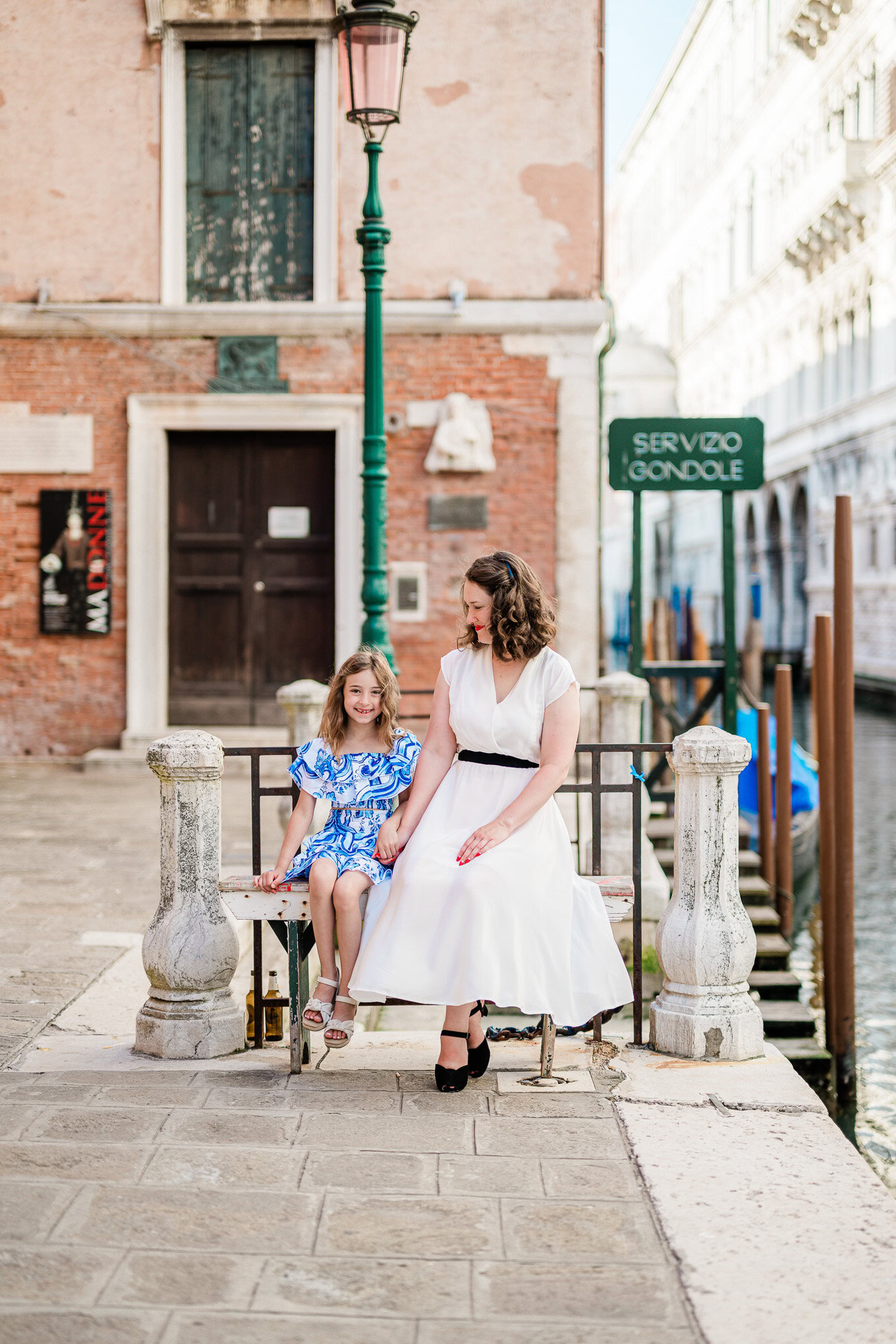 A woman and a girl sit together on a Venice canal bench, enjoying a sunny day with historic buildings and a canal in the.