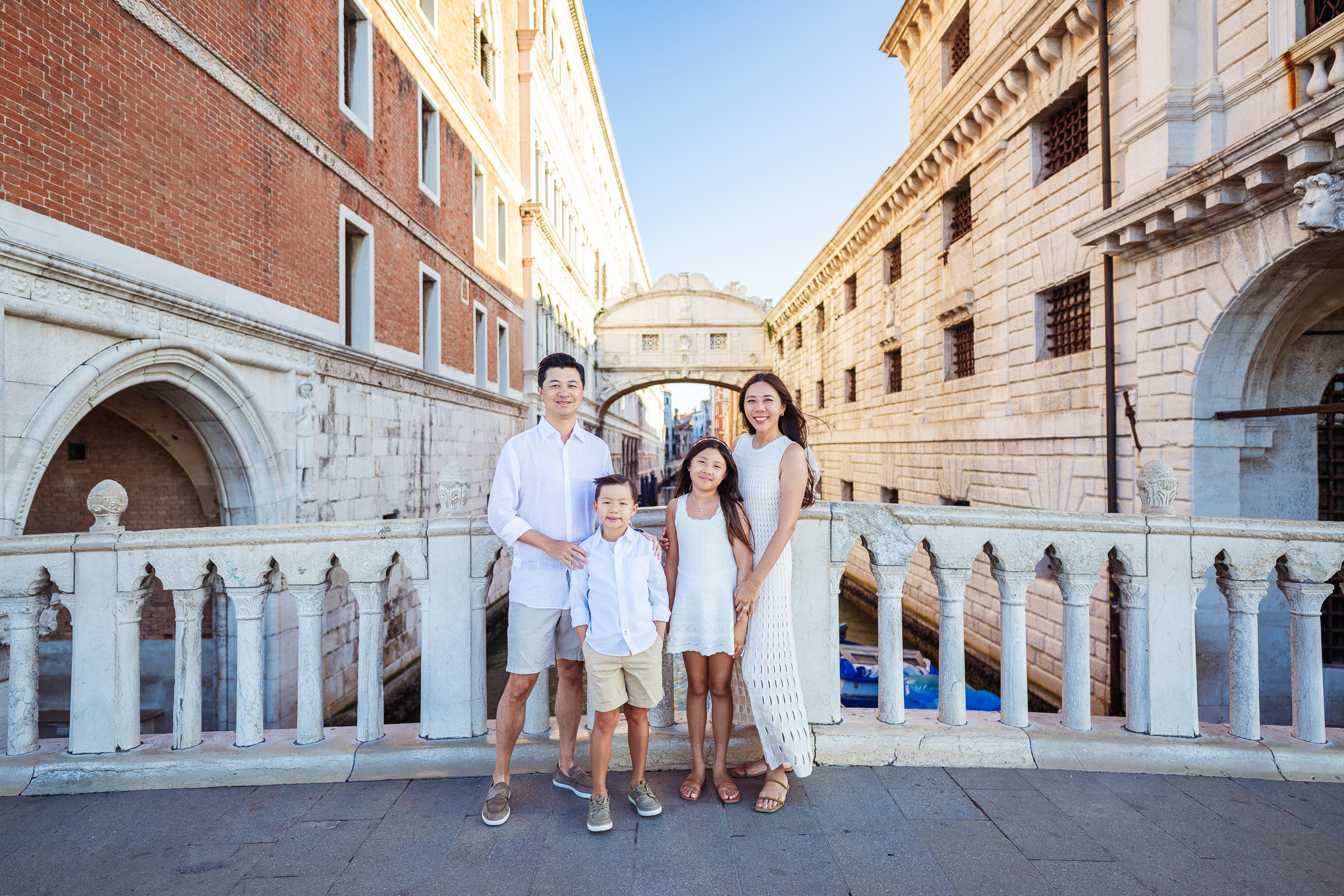 A family of four enjoying a walk along a historic Venetian canal under bright daylight, surrounded by classic architectu.