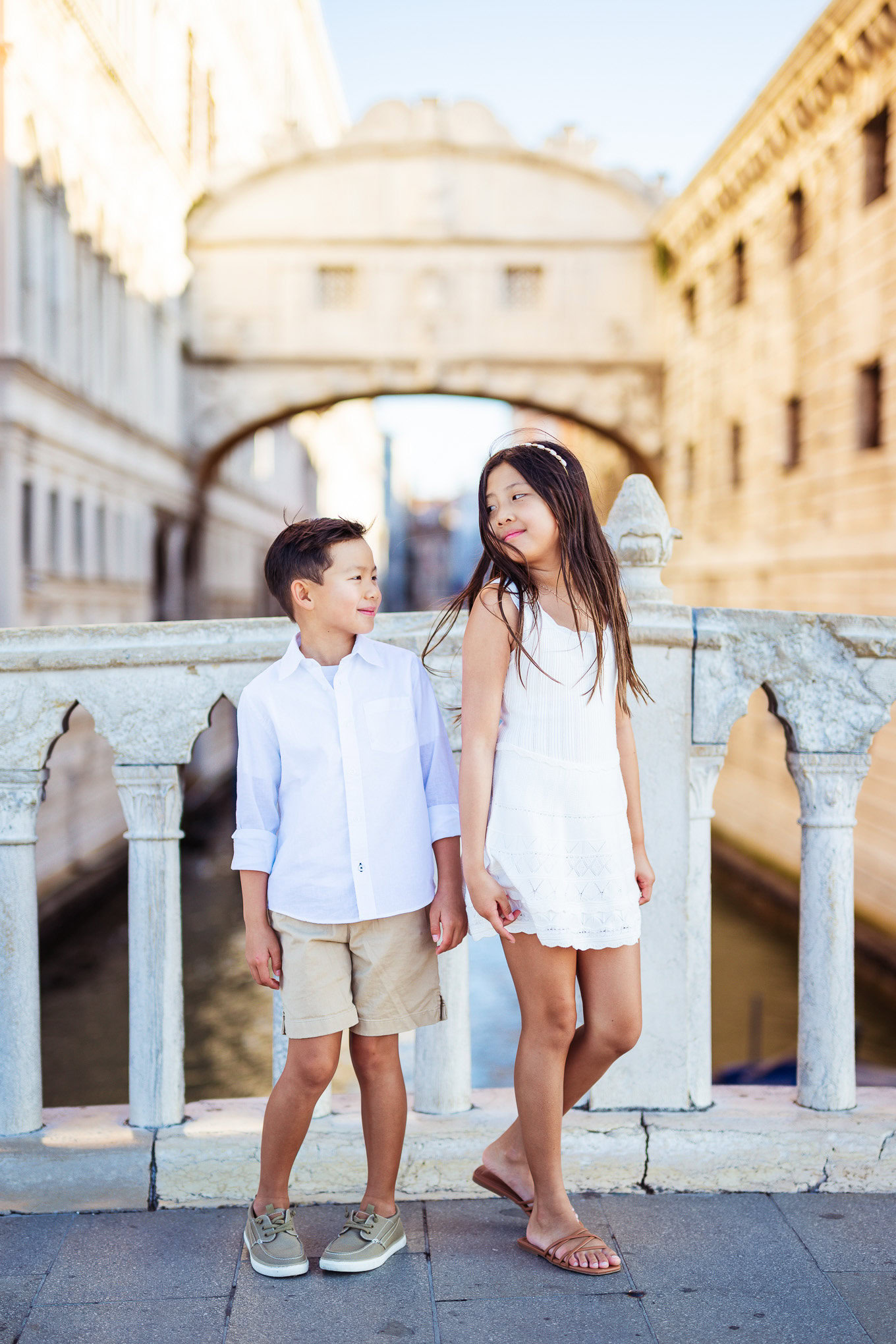 A young girl and boy sharing a joyful moment on a Venetian bridge, illuminated by soft natural light, with historic arch.