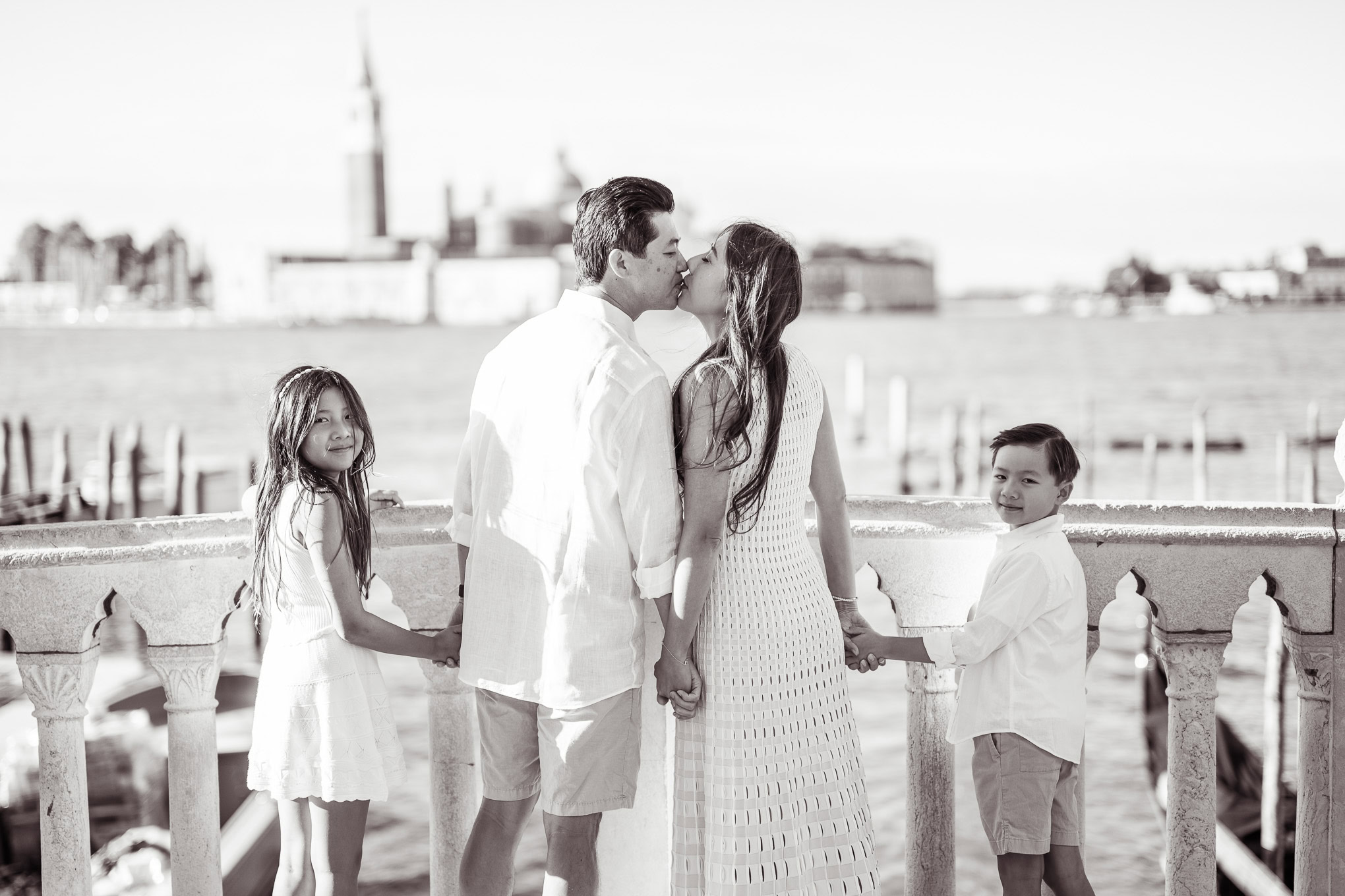 A family holding hands and sharing a tender moment on a Venetian waterfront, with historic buildings and a calm river in.