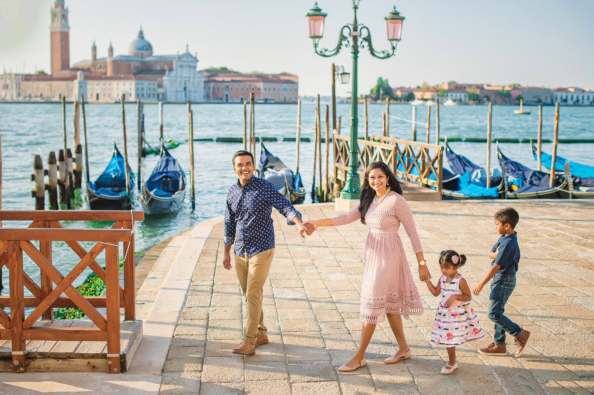 A family holding hands and walking along a Venetian waterfront promenade with boats and historic buildings in the backgr.