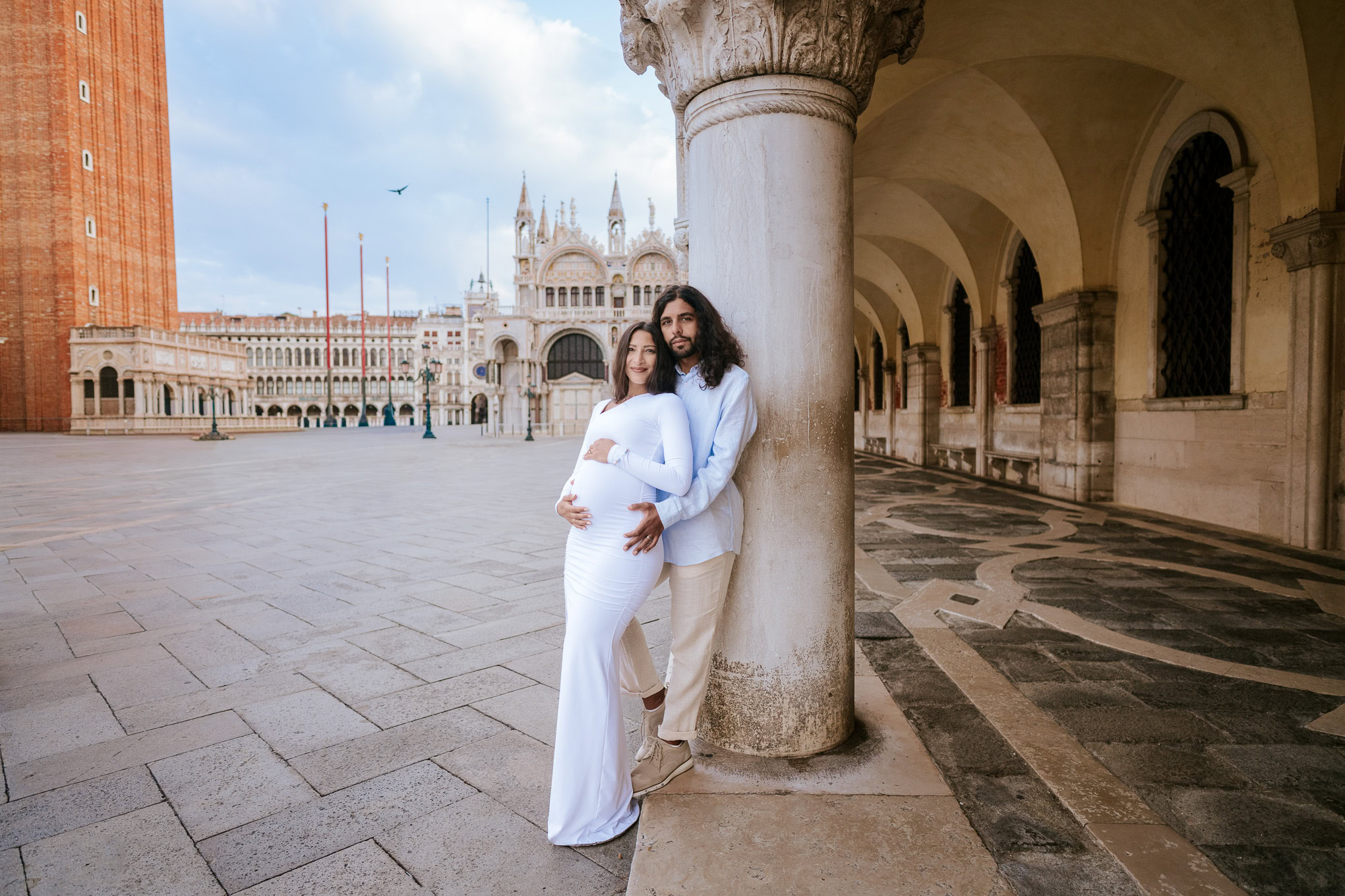 A pregnant couple sharing a tender moment under historic Venetian architecture, bathed in soft natural light.