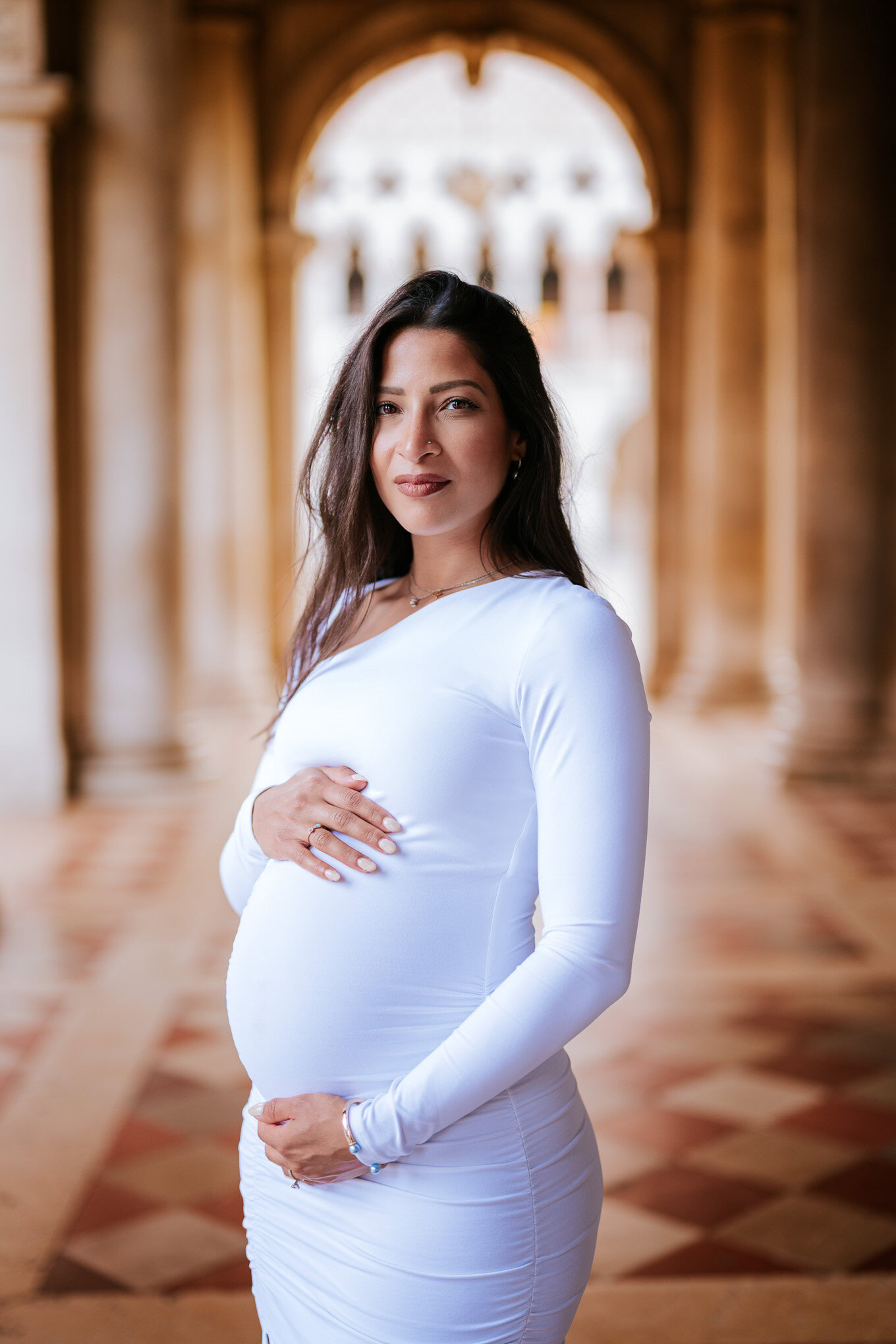 A pregnant woman standing in an ornate Venetian architectural setting with warm lighting and classical details.