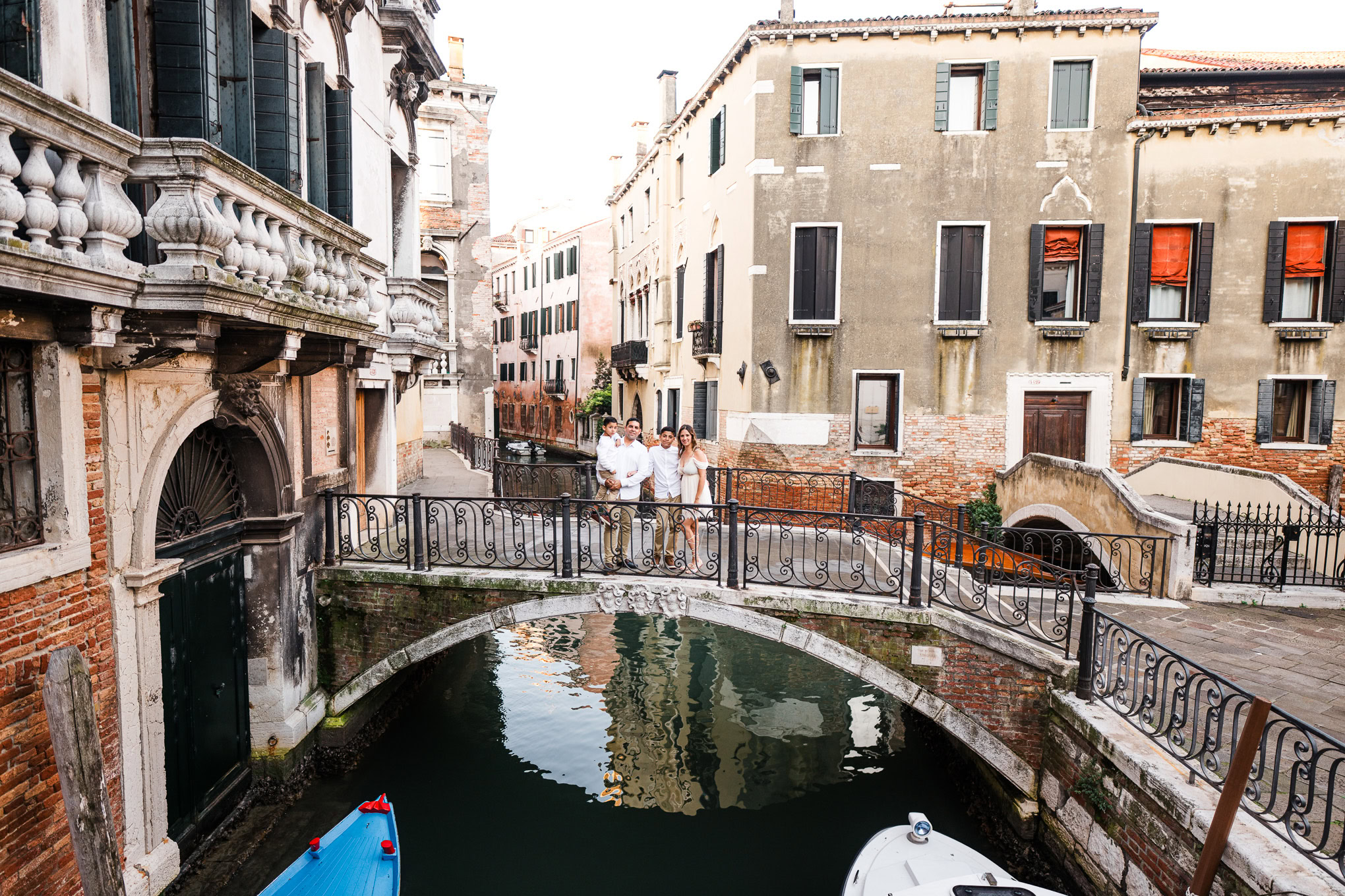A colorful family interacts on a small bridge over a canal in Venice, surrounded by historic buildings and soft natural.
