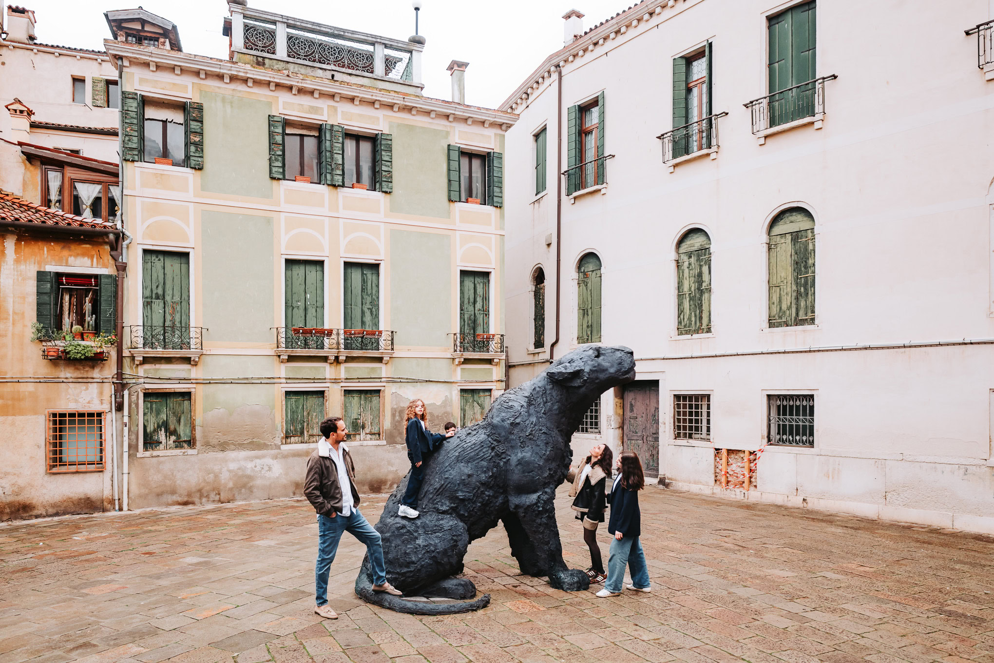 People observing a large dinosaur sculpture in a Venetian courtyard with historic buildings in the background.