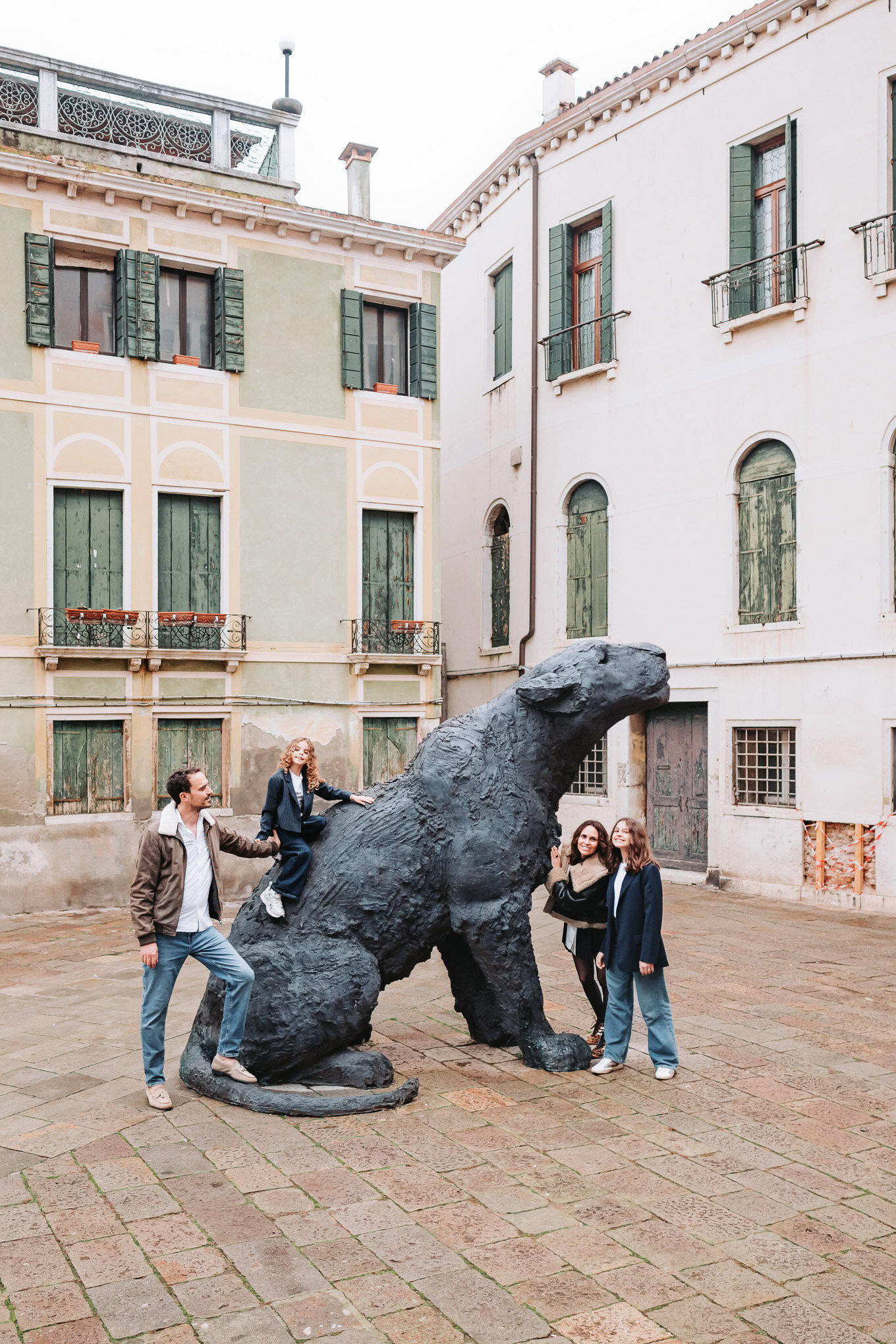 A family interacts playfully around a large dinosaur sculpture in a historic Venetian square, with soft natural light il.