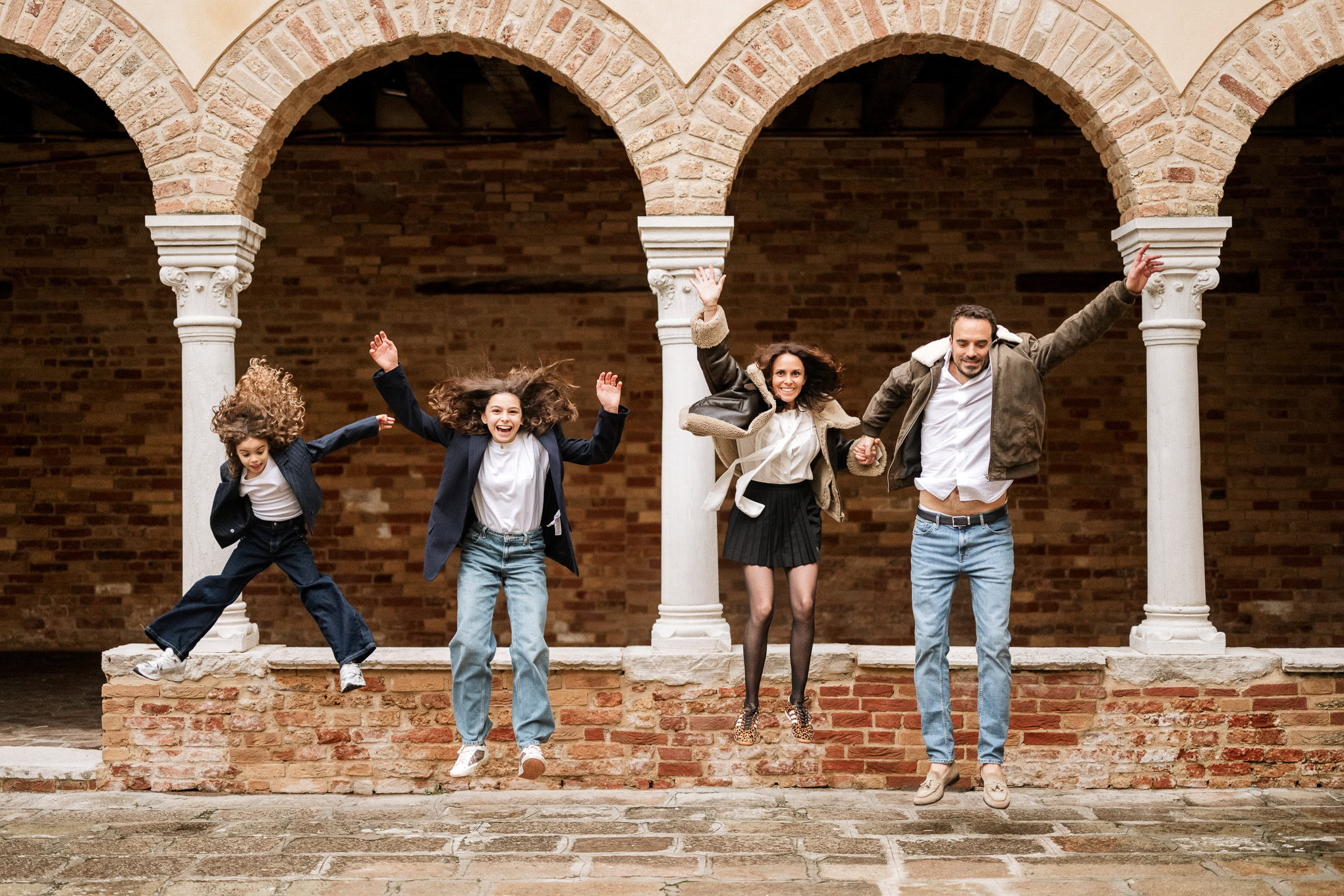 A family of four joyfully jumping and playing together under historic Venetian arches in natural daylight.