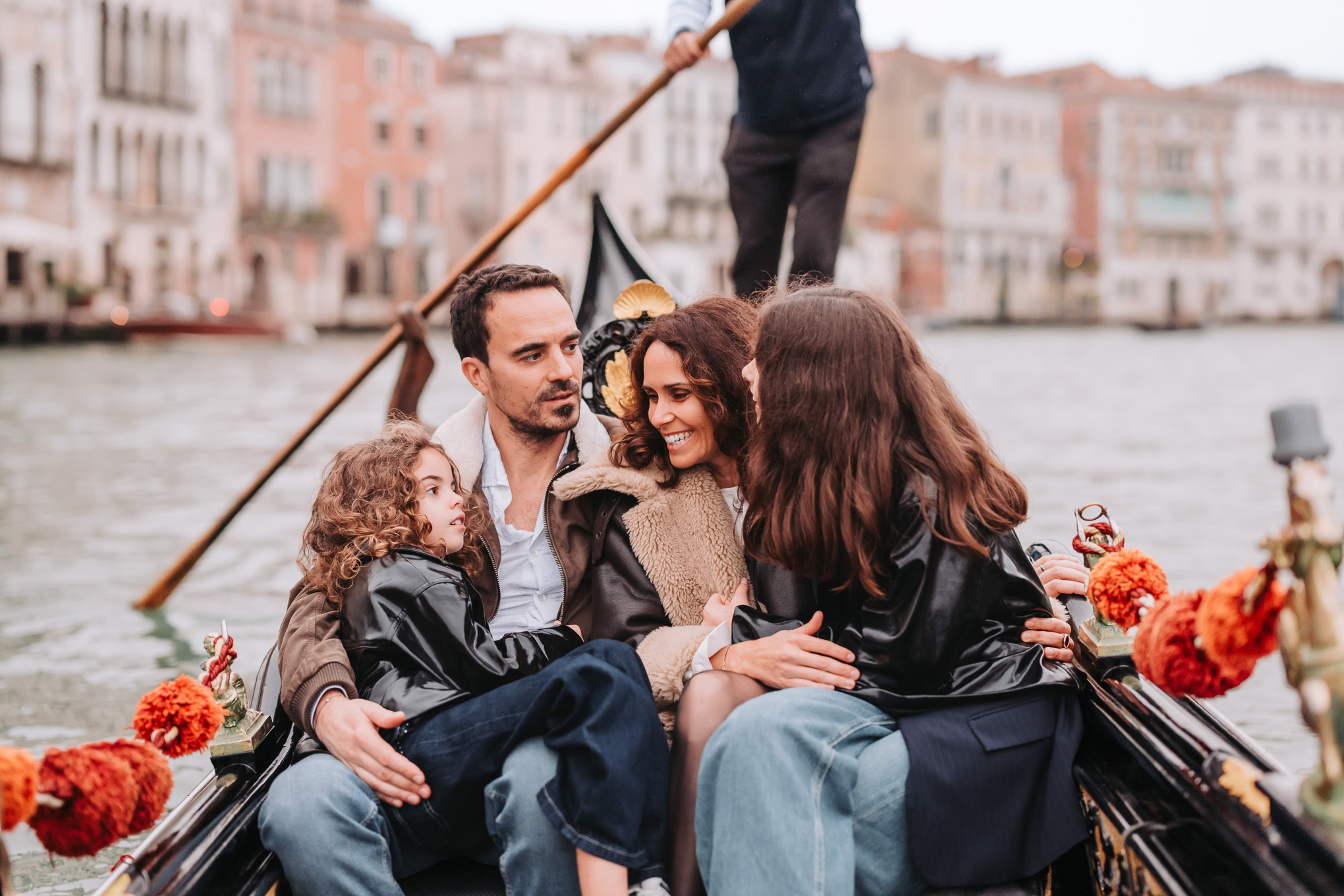 A family enjoying a gondola ride in Venice, engaging in warm conversation amid historic buildings and soft natural light.