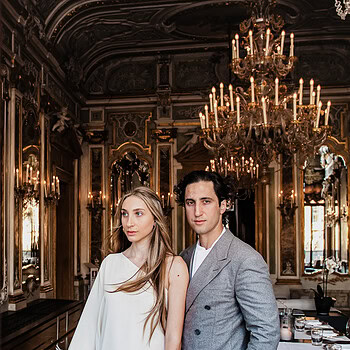 A woman in a white dress with feather details standing in an ornate, chandelier-lit Venetian interior.