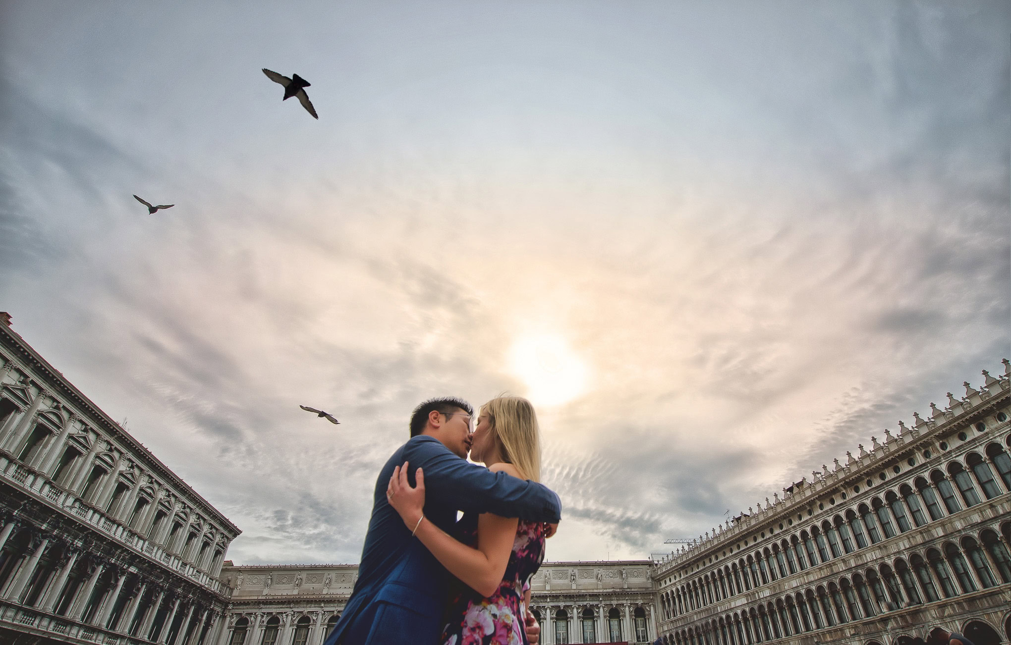 Romantic couple embracing at sunset in Venice's historic square with soft light and flying birds overhead.