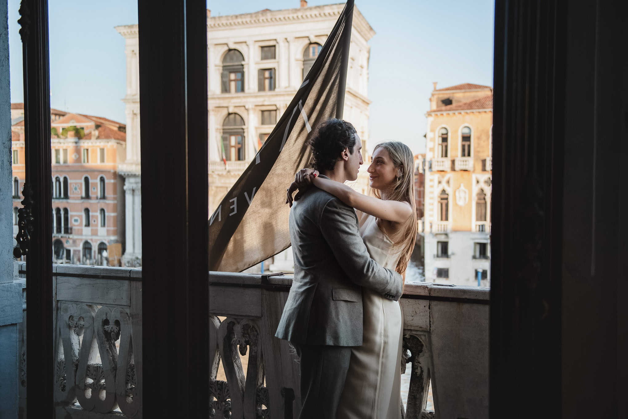 A couple embracing tenderly on a balcony in Venice, bathed in soft natural light, creating a romantic and intimate atmos.