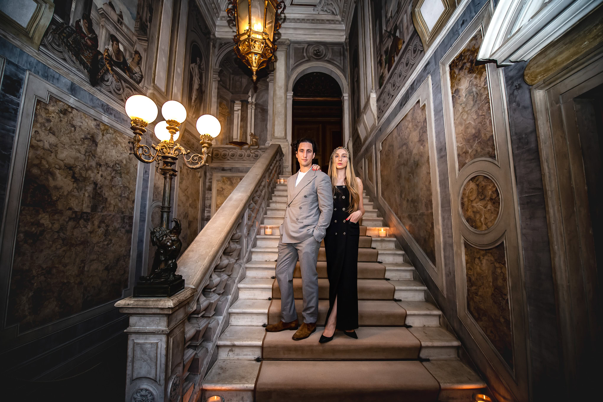 A couple standing on a grand marble staircase inside a luxurious, ornate Venetian palace with warm lighting and elegant.
