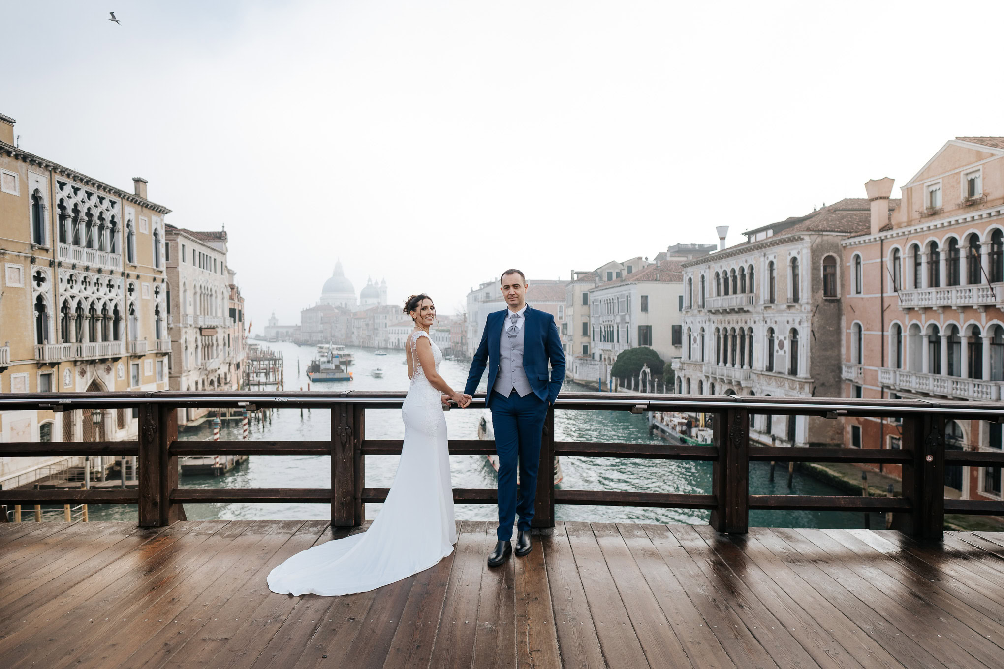 A couple holding hands on a wooden balcony overlooking Venice's canals in soft, romantic morning light.