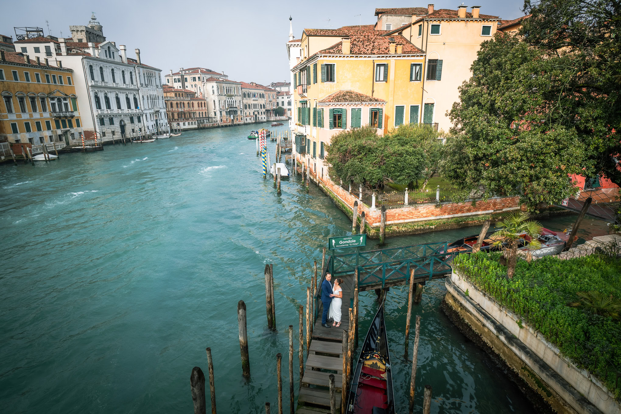 Romantic couple standing on a small bridge over a canal in Venice with warm sunlight and historic colorful buildings.
