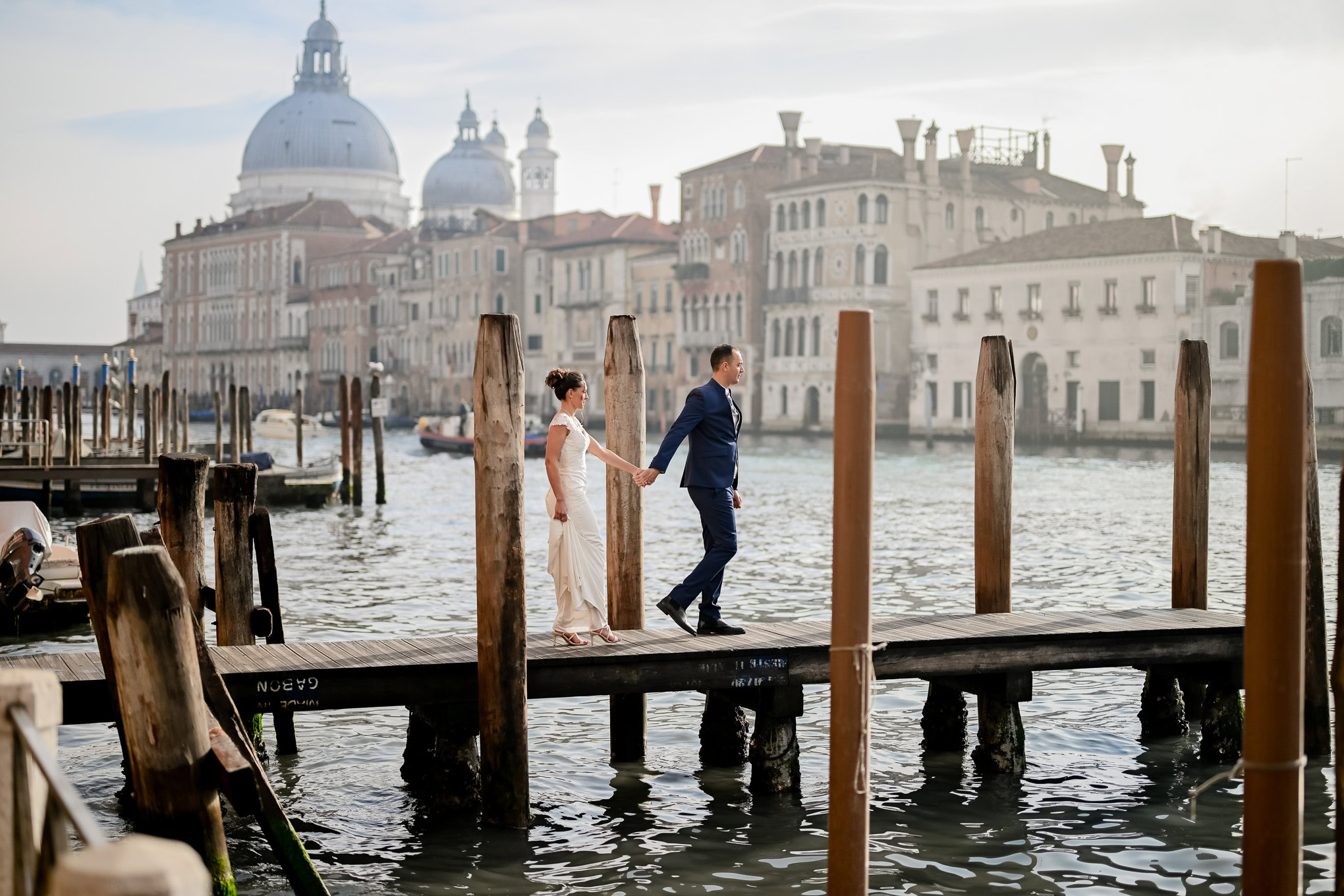 A woman and man walking hand in hand along a Venetian dock with historic buildings and domes in the background.